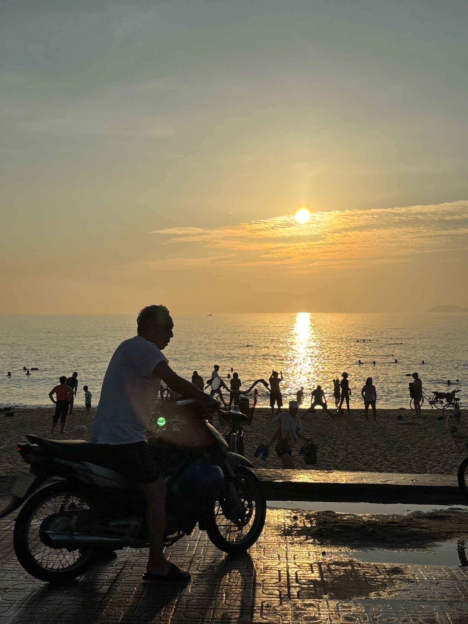 A silhouetted man sits on a motorcycle in the foreground, facing right, with the ocean and a beach in the background. The setting sun casts a warm, golden glow over the scene, reflecting brightly on the water. Several figures, also silhouetted, are visible on the beach, some wading in the shallow water. The beach appears to be a public area, with people engaged in various activities, suggesting a communal gathering or a public park by the sea. The overall impression is one of a serene evening by the coast. The presence of a motorcycle suggests proximity to a road or pathway adjacent to the beach. This setting is characteristic of a coastal city like Nha Trang, Vietnam, known for its beautiful beaches and vibrant street life.