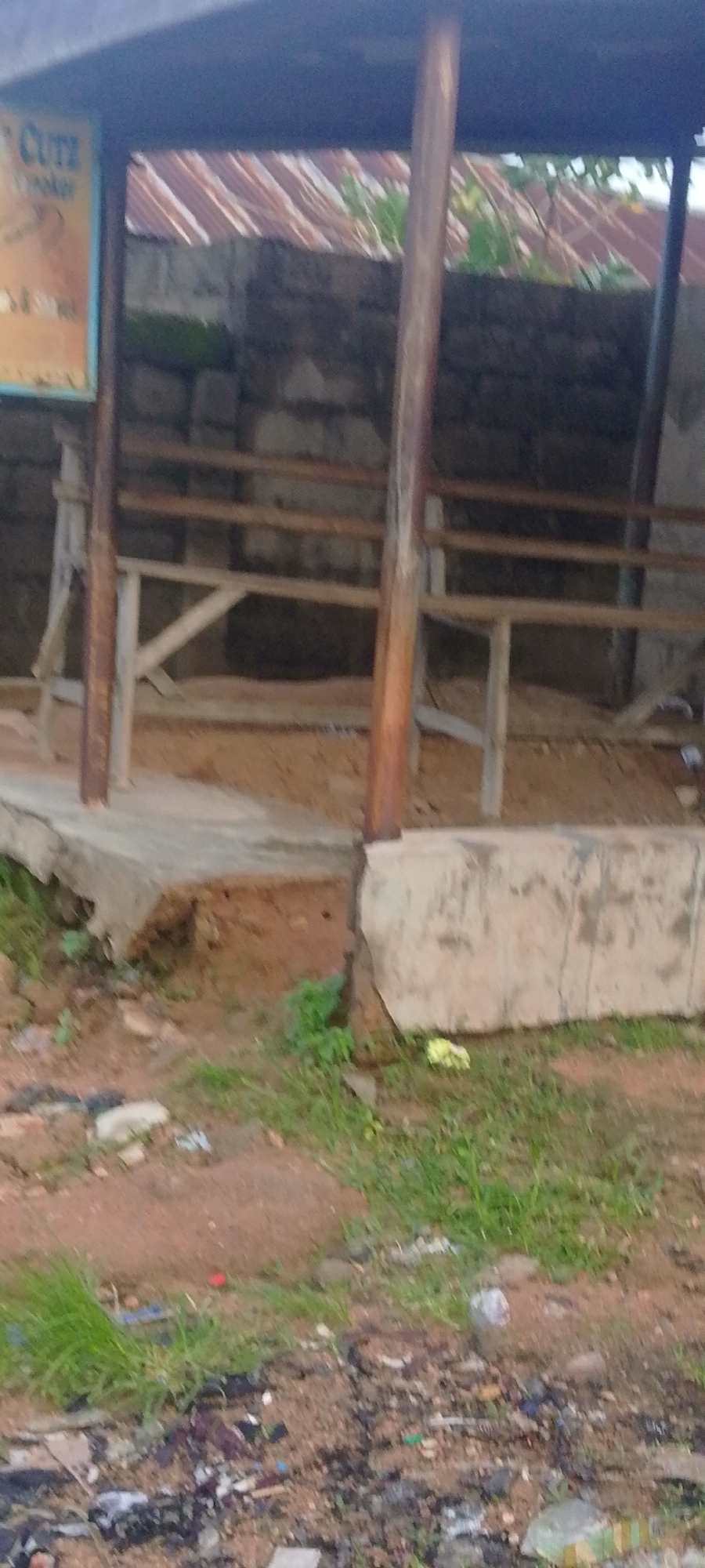 The image shows a rustic, open-sided structure, possibly a shelter or market stall, constructed with wooden posts and a corrugated metal roof. The structure is situated on a concrete platform that appears weathered and cracked, with exposed earth and some vegetation growing around its base. Behind the shelter, a rough stone wall is visible, topped with more corrugated metal sheeting, suggesting the presence of an adjacent building or enclosure. The ground in front of the structure is uneven, composed of dirt and scattered debris, including plastic waste and dry leaves. In the foreground, the ground is covered with more debris and sparse green grass. The overall impression is of an unfinished or neglected outdoor space in an area that experiences varying weather conditions, consistent with a location like Jalingo, Nigeria, where such informal structures are common. There are no people or animals visible in the frame.