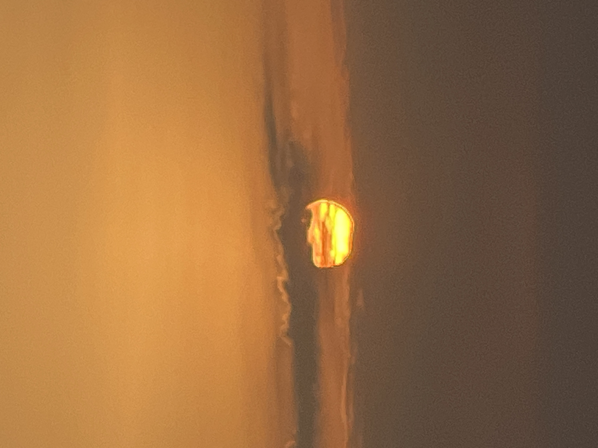 The image shows a sunset over the ocean in Kuta, Indonesia. The sun is a vibrant orange disc partially obscured by clouds, casting a warm glow across the sky. The water below is a hazy, muted brown, indicating the presence of sand or sediment, typical of the Kuta coastline. There are no people or man-made objects visible in the frame. The focus is entirely on the natural elements of the seascape.
