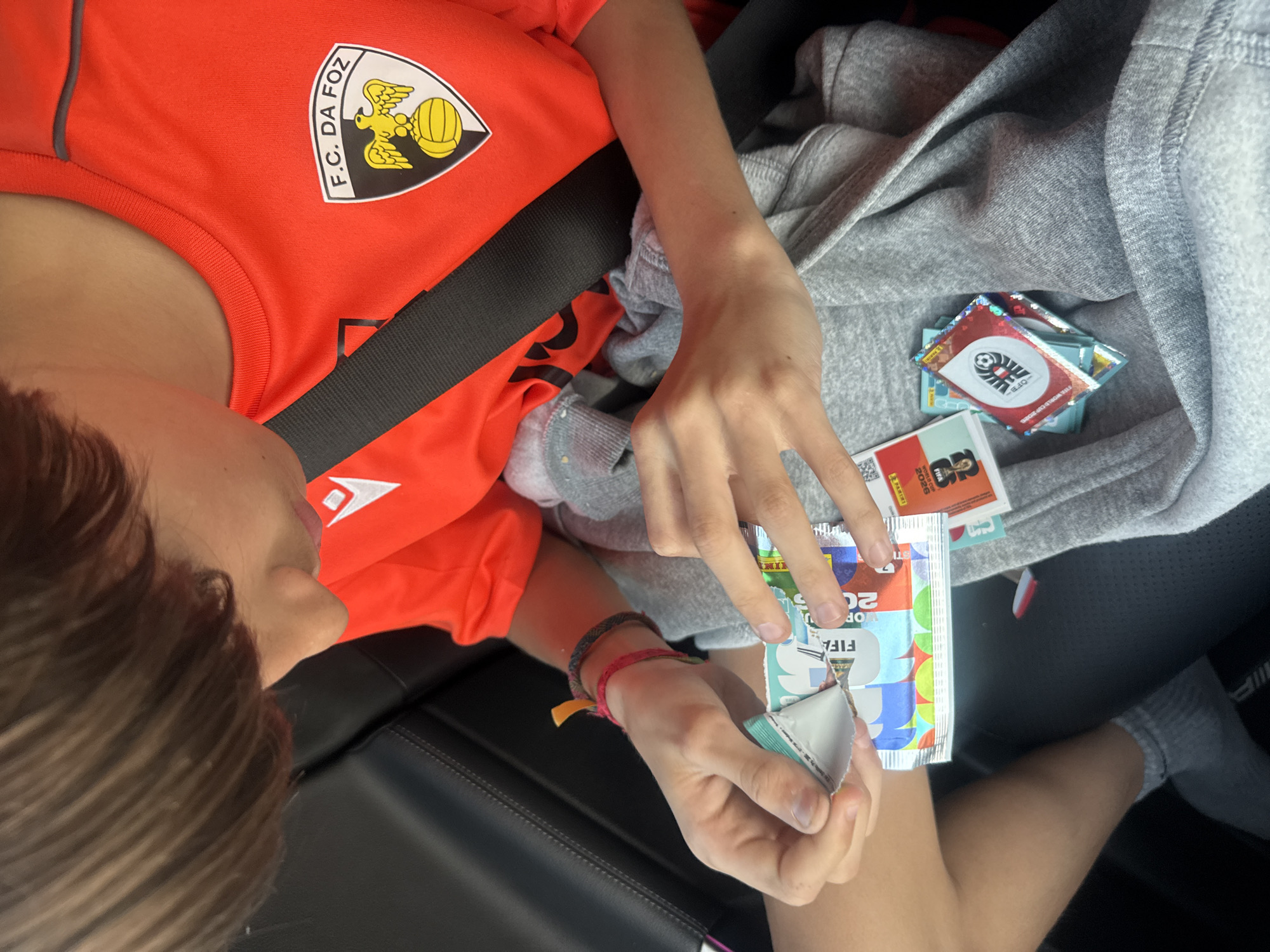 A child in an orange F.C. Da Foz soccer jersey with a seatbelt on is opening FIFA World Cup 2026 Panini sticker packs while seated in a vehicle. The vehicle appears to be black with grey seats. The child is surrounded by opened sticker packs and loose stickers. The picture was taken inside a car somewhere in Vila Nova de Gaia, Portugal.