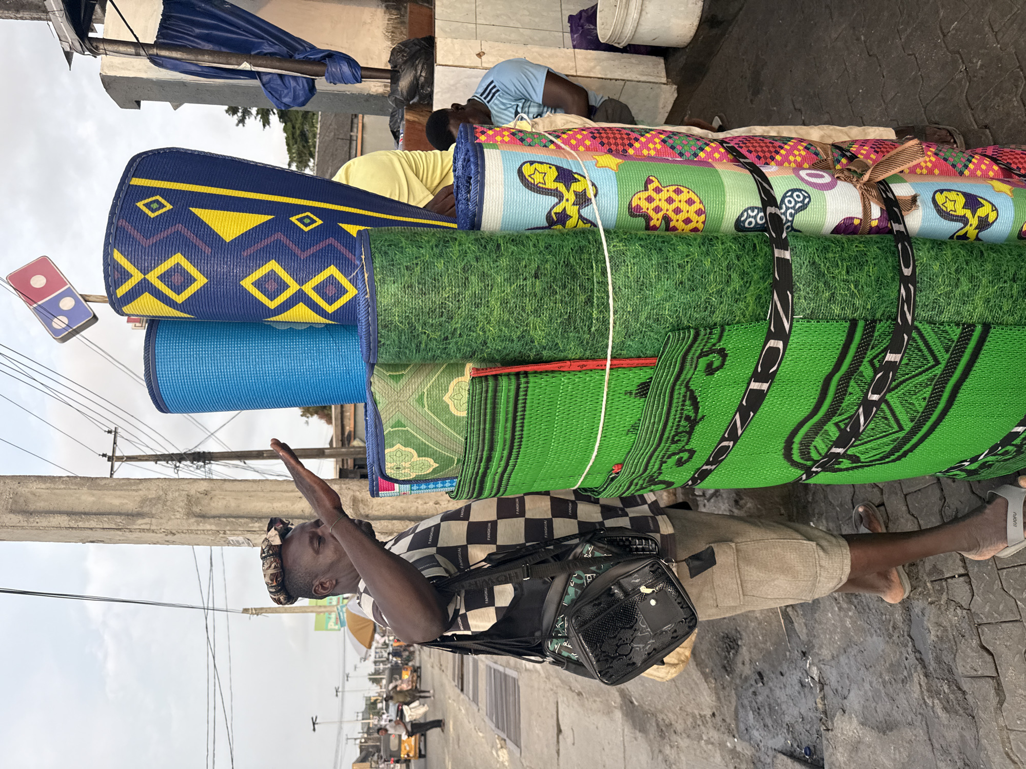 In Ado, Nigeria, a man wearing a checkered shirt and khaki shorts stands on a paved sidewalk next to a towering stack of rolled-up mats. He is holding his right hand up to his forehead as if shielding his eyes from the sun. A black messenger bag with a patterned design hangs from his left shoulder. The mats, made of brightly colored plastic, are bound together by ropes and straps, displaying various patterns, including geometric designs, images of grass, and cartoonish figures. In the background, a Domino's Pizza sign is visible, along with other street scenes, including vehicles and pedestrians. The sky is overcast, suggesting a cloudy day.