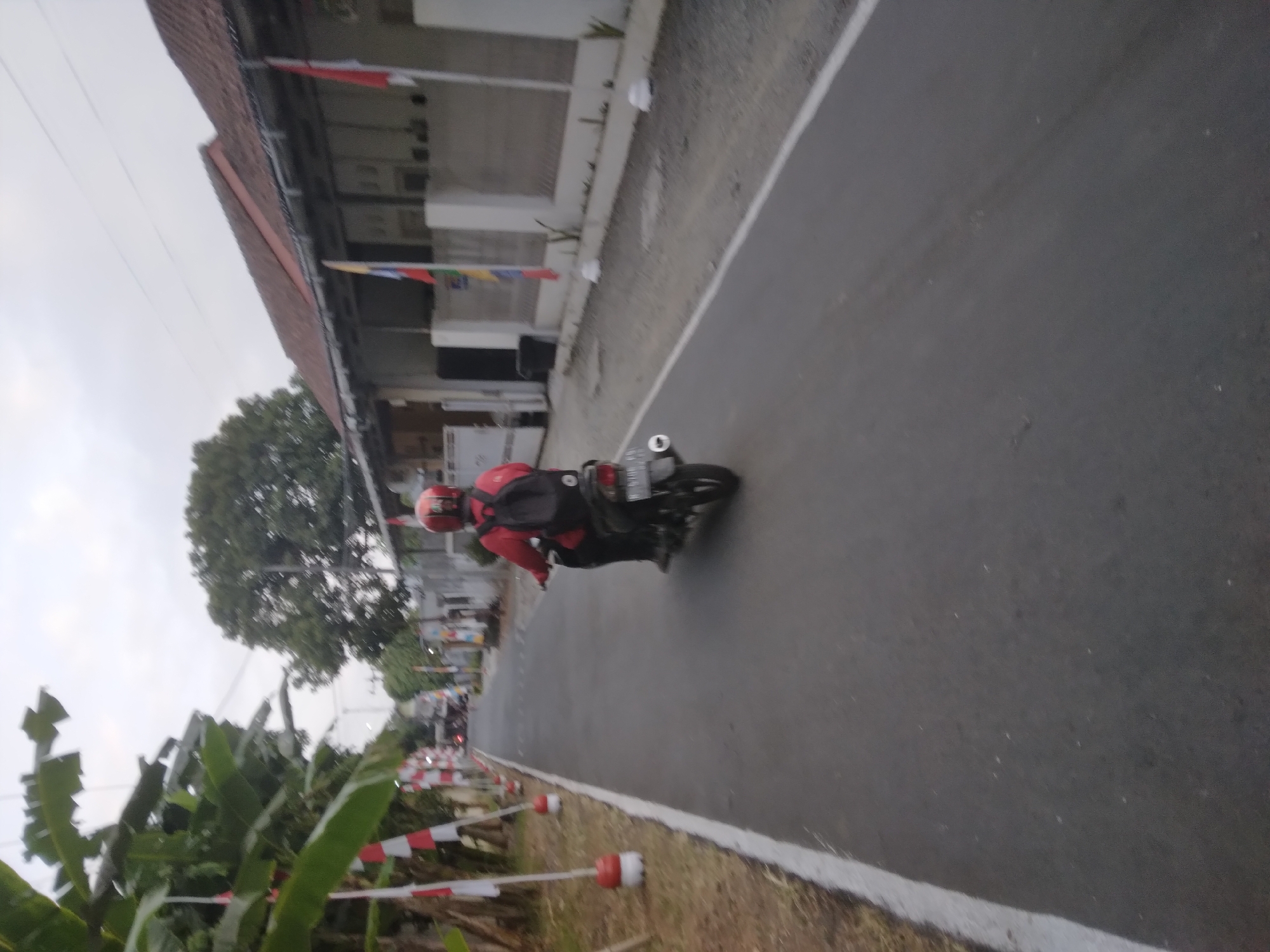 In Desa Pamijen, Kecamatan Sokaraja, Indonesia, a person wearing a red helmet and jacket rides a black motorcycle down an asphalt road. The rider has a black backpack. The road is lined with white markings on the left and a grassy area with red and white flags on poles on the right side of the road. Behind the flags, houses with red-tiled roofs are visible, and some also have flags displayed. The sky is overcast, and the overall lighting suggests it is either early morning or late afternoon.