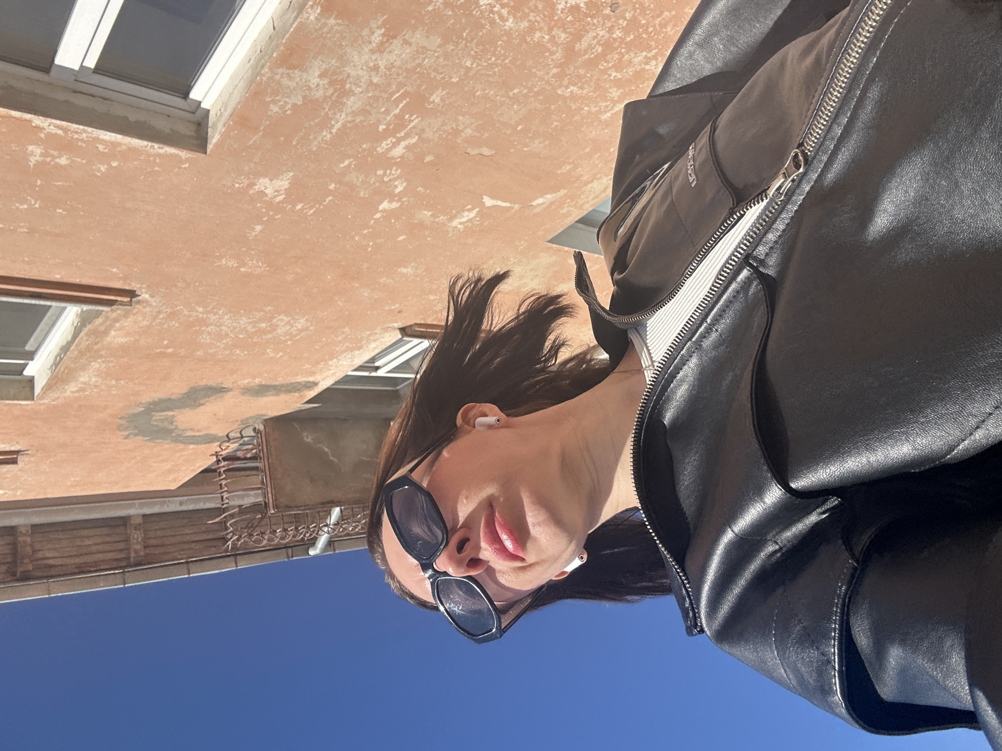 A low-angle, rotated photograph shows a person wearing a black leather jacket and sunglasses, standing beneath a building in Votkinsk, Russia. The person has short, dark hair and appears to be wearing wireless earbuds. A building with a faded orange facade and white-framed windows is visible overhead, and a section of rusted metalwork is attached. The background shows a clear blue sky.