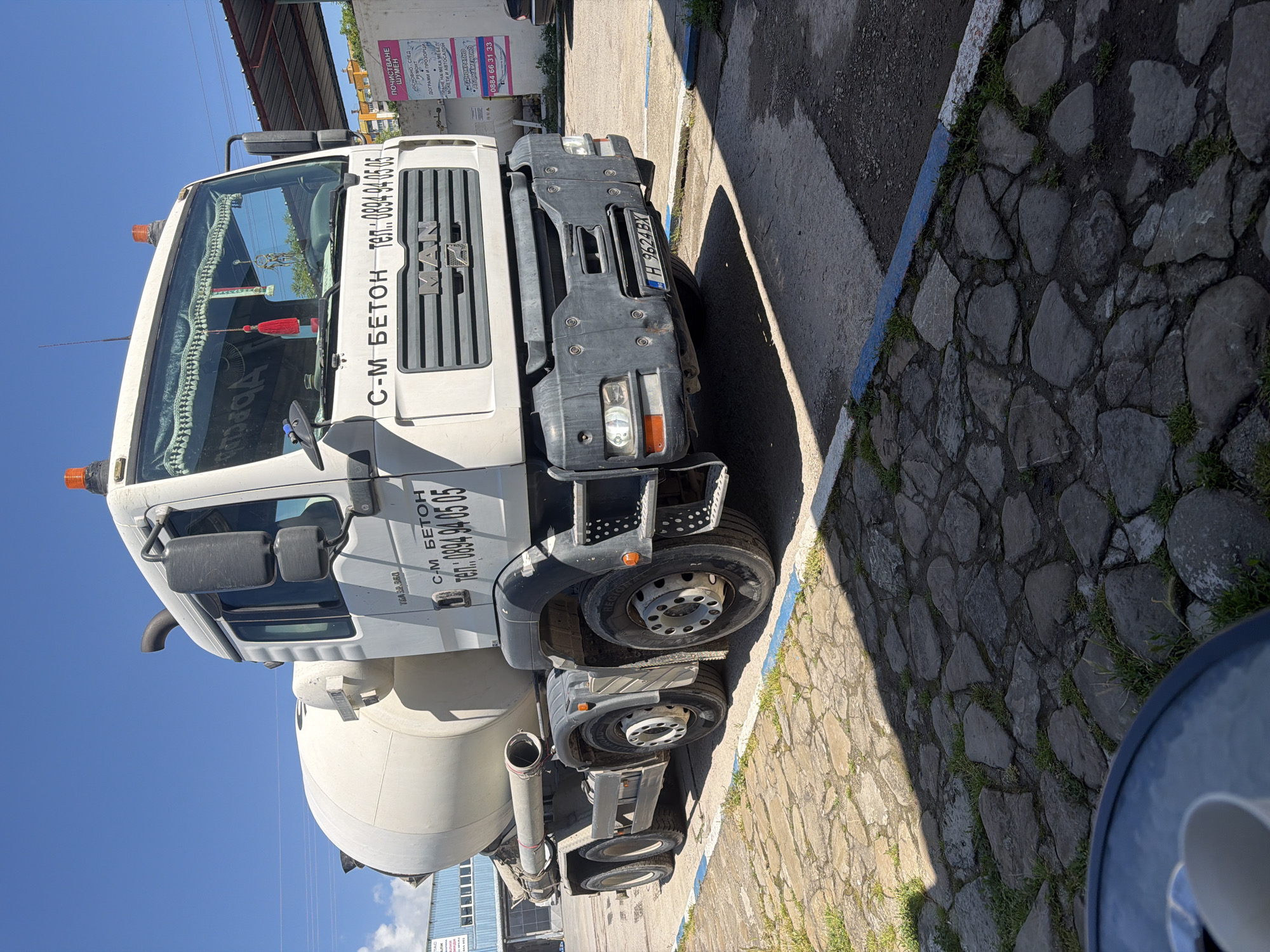 A white MAN cement mixer truck is the central subject of this image, which is rotated 90 degrees clockwise. The truck is parked outdoors on a sunny day, evident from the clear blue sky and distinct shadows cast on the ground.
The vehicle is white with a grey bumper and steps, and it features orange warning lights on its roof. On the truck's cabin, the text "С-М БЕТОН" (S-M BETON) and the phone number "тел: 0894 94 05 05" are visible. The manufacturer's name, "MAN," is on the front grille. The license plate "H 9624 BX" is affixed to the front bumper, indicating its registration in Shumen, Bulgaria.
The truck is situated on a paved surface made of irregularly shaped stones with grass growing in the crevices, next to an asphalt patch. A blue painted line runs along the edge of the stone paving. In the background, under a bright sky with scattered clouds, parts of a building are visible, along with a white sign displaying information in Cyrillic. The sign advertises "ПОЧИСТВАНЕ ШУМЕН" (Cleaning Shumen), listing services like "ОСНОВНО СЛЕД МОНТАЖ" (Deep cleaning after installation), "ДОГРАМИ И ПРОЗОРЦИ" (Window frames and windows), "ПРАНЕ НА МЕКА МЕБЕЛ, МОКЕТИ И АВТОСАЛОН" (Washing upholstered furniture, carpets and car interiors), with options for "ЕДНОКРАТНО" (One-time) or "АБОНАМЕНТНО" (Subscription) services, and a contact number "0884 66 31 33".
No people are visible in the image. The scene appears to be an industrial or service area in Shumen, Bulgaria, during daytime.