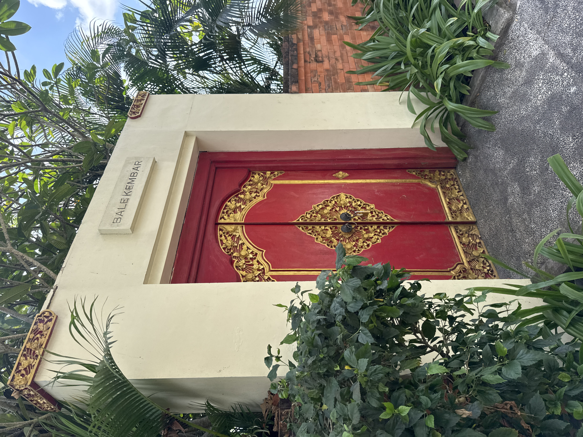 A traditional Balinese entryway in Kuta Selatan, Indonesia, features a striking bright red double door adorned with ornate gold carvings. Above the door, a stone plaque displays the text "BALE KEMBAR" in capital letters. The entryway is framed by a cream-colored wall, topped with decorative gold trim, and lush green foliage, including palm fronds, spills over from above. To the right of the door, a rustic red brick wall is visible, bordered by vibrant green plants. The ground is paved with dark speckled stone. The scene is illuminated by natural daylight, suggesting it is daytime.