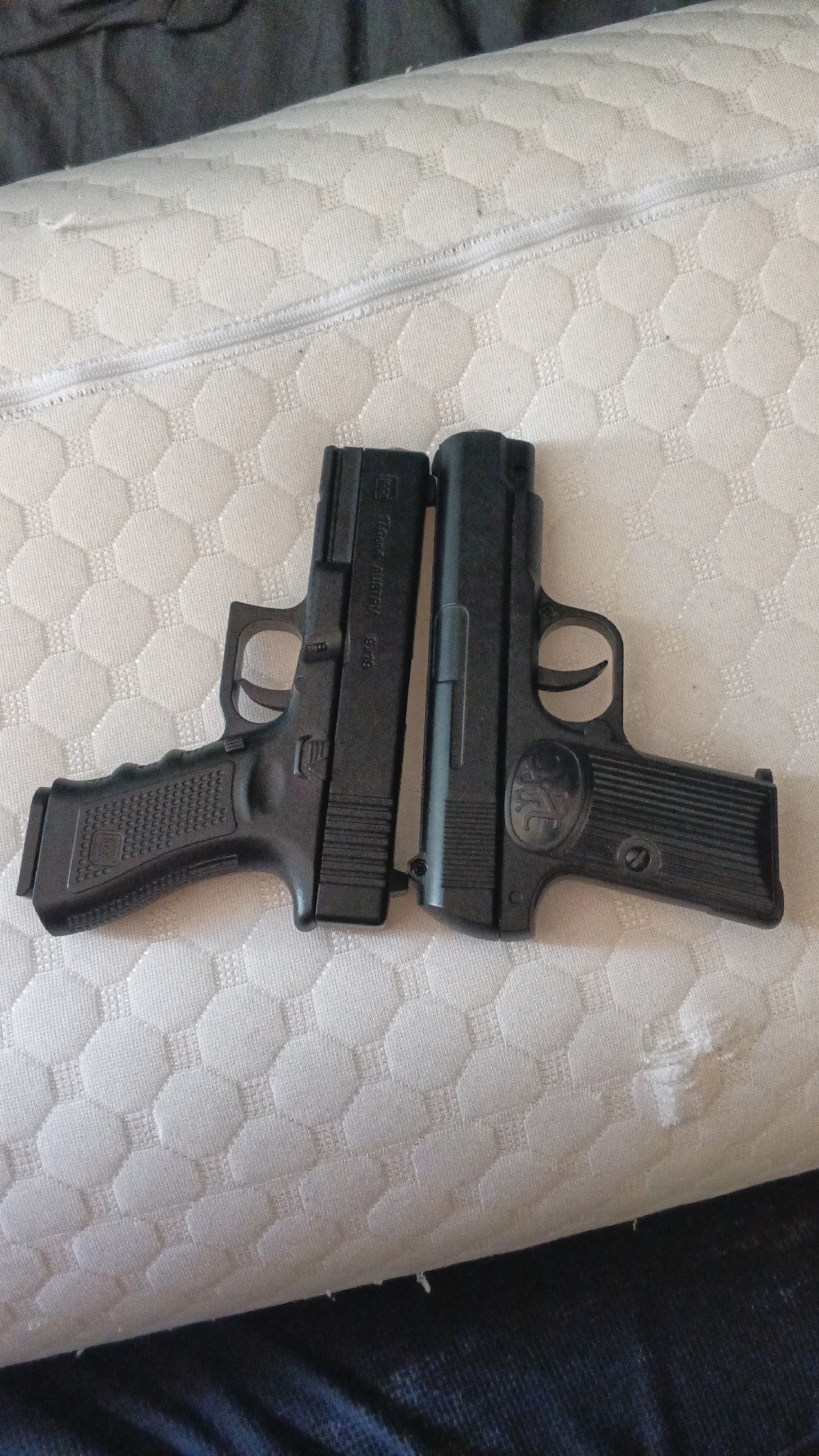 Two black toy guns are placed side-by-side on a white textured mattress. The gun on the left is a replica of a Glock pistol, with "Glock Austria 9x19" inscribed on its slide. The gun on the right is a replica of a Browning pistol, with "FN" logo on its grip. The guns are oriented horizontally, with their barrels pointing to the right. The background is a dark fabric, possibly a bedsheet or blanket. The image is taken from a top-down perspective. There is no information to suggest the location is in Sochi, Russia.