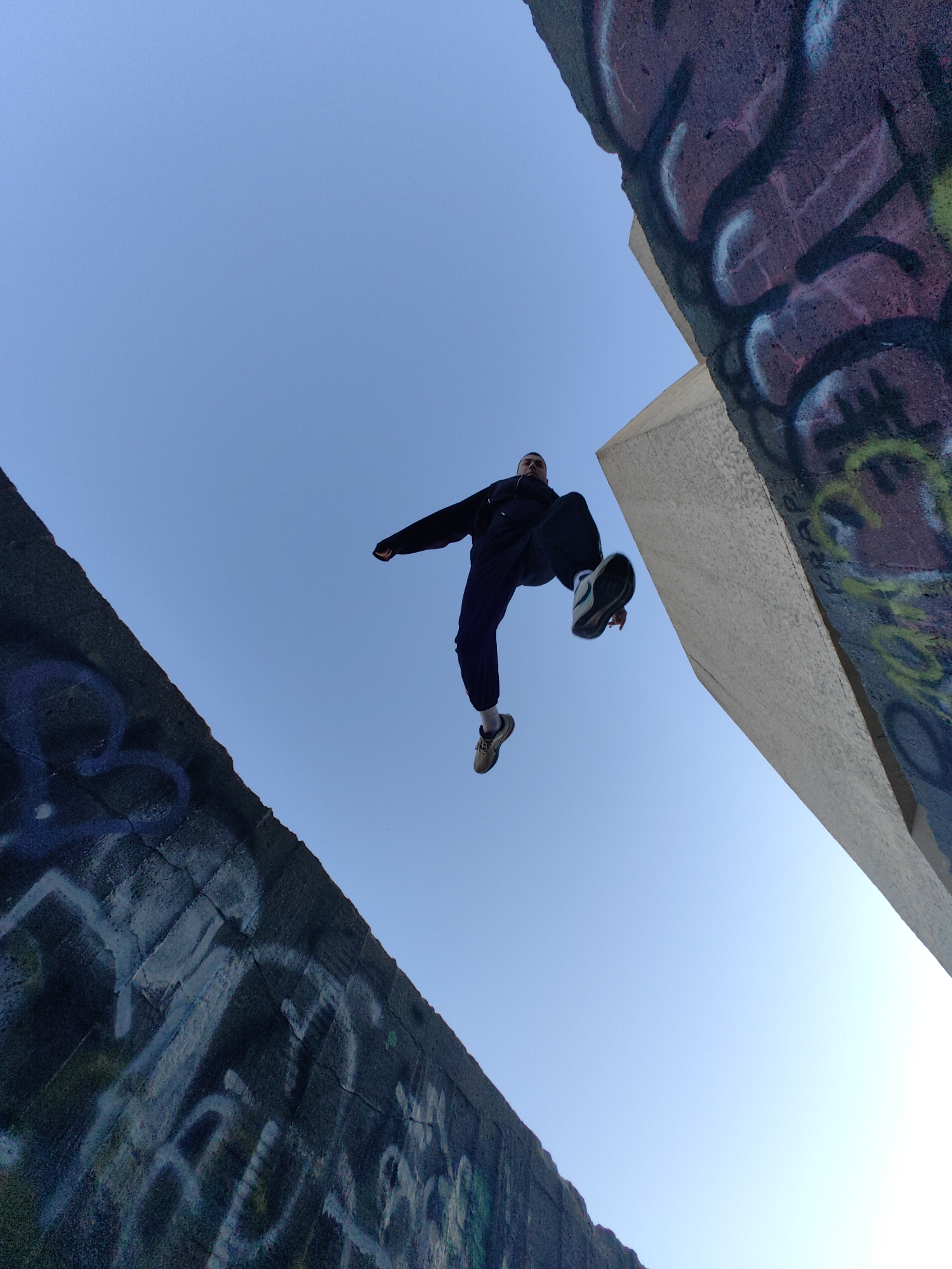 From a low-angle perspective, the image captures a person mid-jump, appearing to leap between two graffiti-covered concrete structures against a clear blue sky. The person is dressed in dark clothing. The structures are heavily marked with graffiti. The location can be identified as Varna, Bulgaria, from the style and materials used in the construction, as well as the nature of the graffiti present.