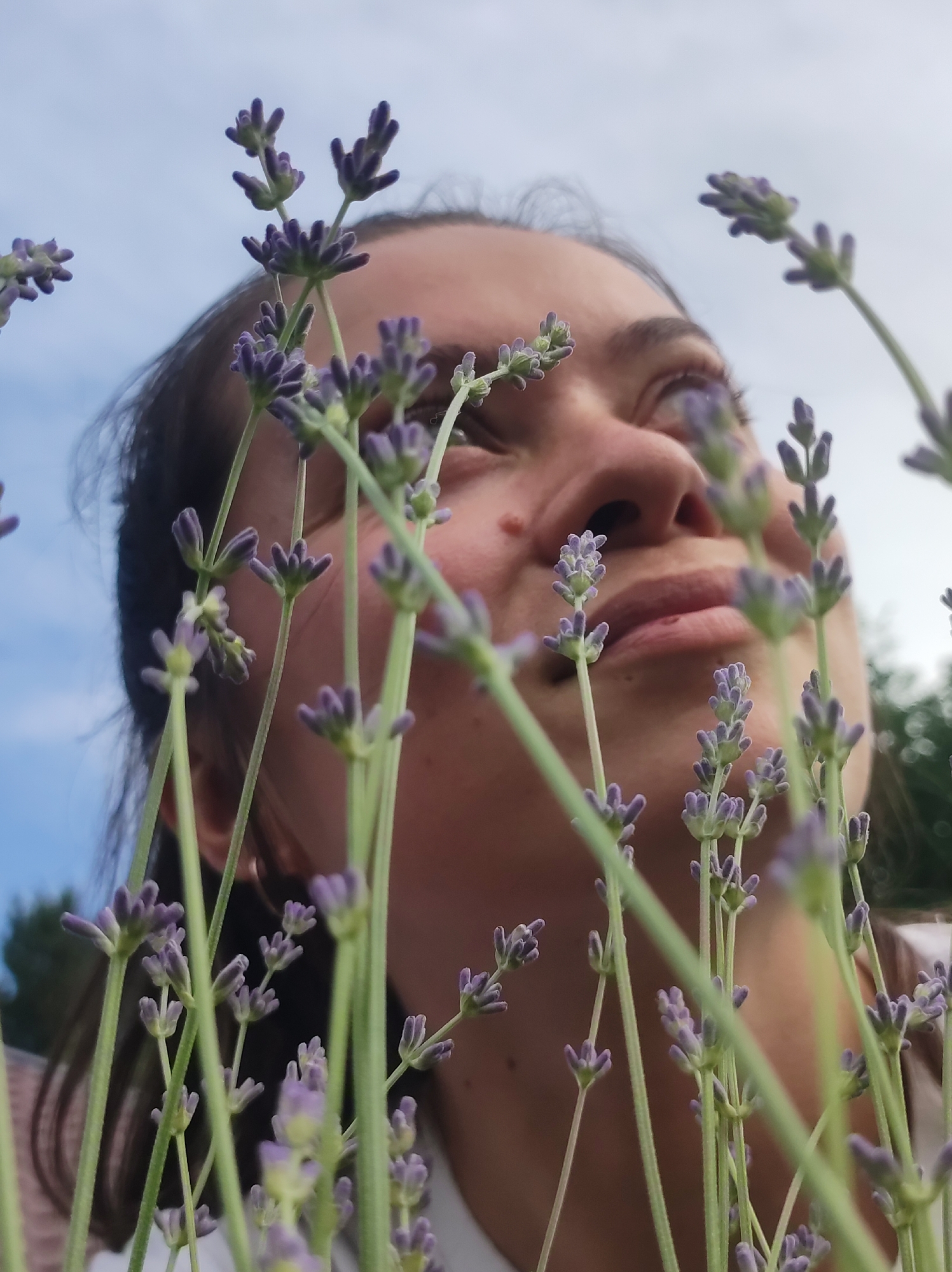 A low-angle, medium shot captures a person looking upwards through a field of lavender flowers. The person's face is mostly out of focus, with only the lower portion of their face, including their lips and nose, clearly visible in the center of the frame. Their skin is light-toned with some freckles on the nose. The lavender stalks and purple buds are in sharp focus, obscuring parts of the person's face and creating a natural framing effect. The background consists of a slightly overcast sky and blurred green foliage. The image appears to have been taken outdoors, likely in a garden or a field. No specific location in Фоки, Russia, can be identified from the visual information provided.
