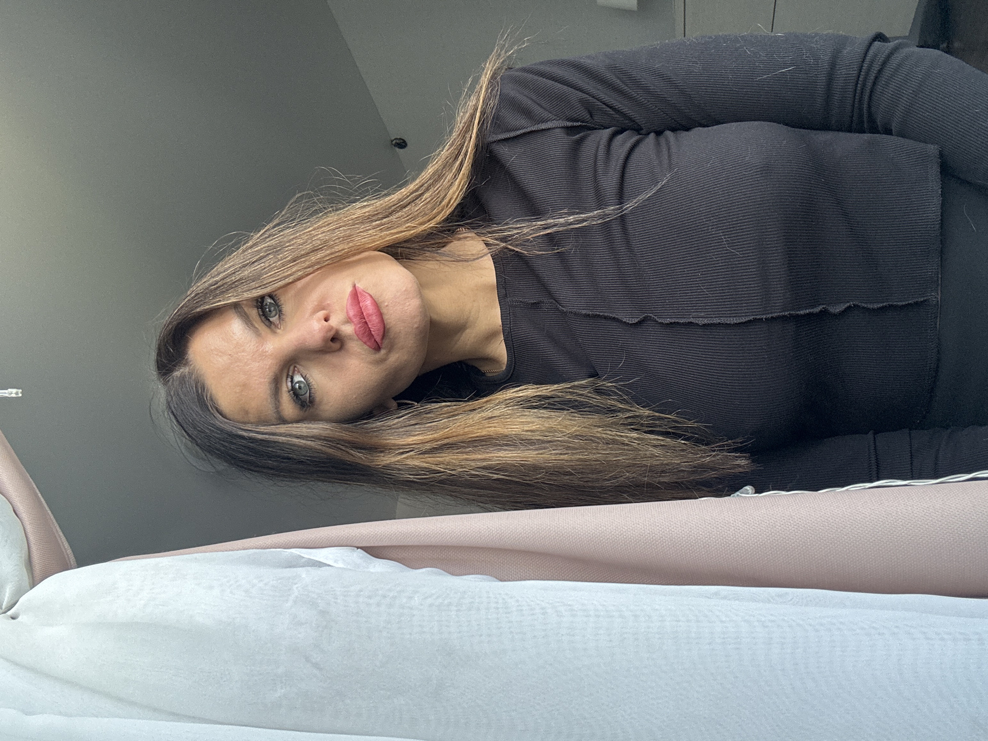 A woman is looking down into the camera. She has long brown and blonde hair, light eyes, and red lipstick. She is wearing a black top. Behind her, there is a wall. In the foreground, there are layers of white, light pink, and darker pink fabric. The location of the image is unknown, and it is not possible to determine if it was taken in the city of Chaikovsky, Russia.