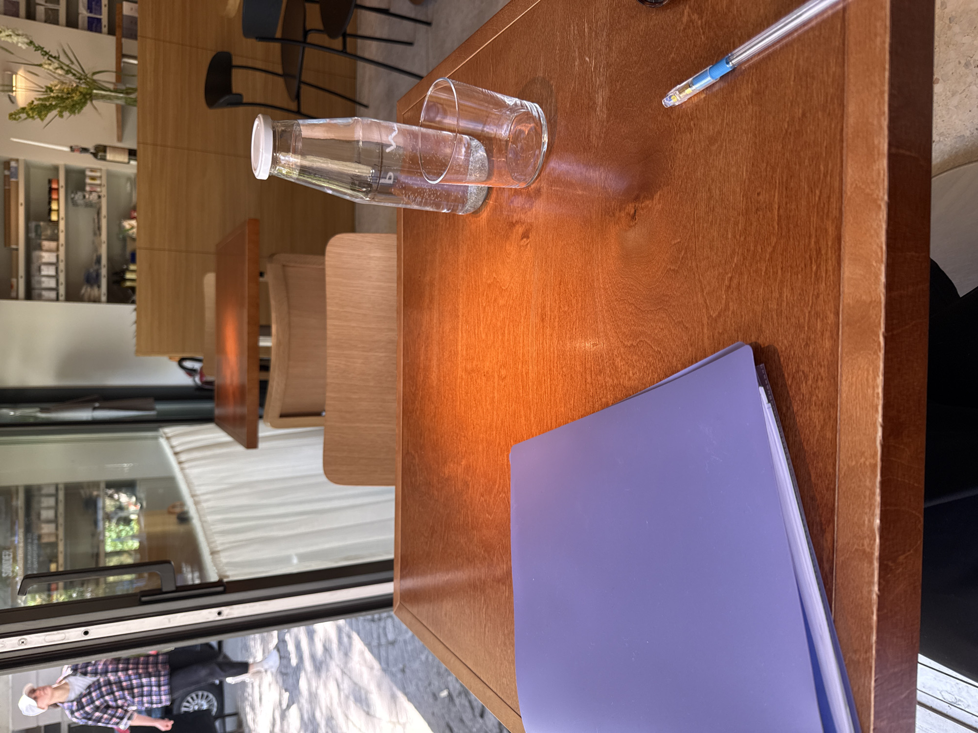 A medium shot, captured from a slightly high-angle perspective, shows the corner of a wooden table in what appears to be a café or restaurant in Belgrade, Serbia.
On the table, near the right edge, lies a blue file folder. Adjacent to it are a half-full glass water bottle and a clear glass cup, both placed horizontally on the table. Further up the table, a pen rests.
In the background, beyond the table, are matching wooden chairs lined up in a row, and behind them, a glass window through which a woman wearing a white cap, a plaid shirt, and dark pants can be seen standing outside near a parked car. Shelves filled with various products, possibly wine or groceries, can be seen in the upper left background of the image. Fresh flowers in a vase are on the top shelf. The scene is naturally lit.
