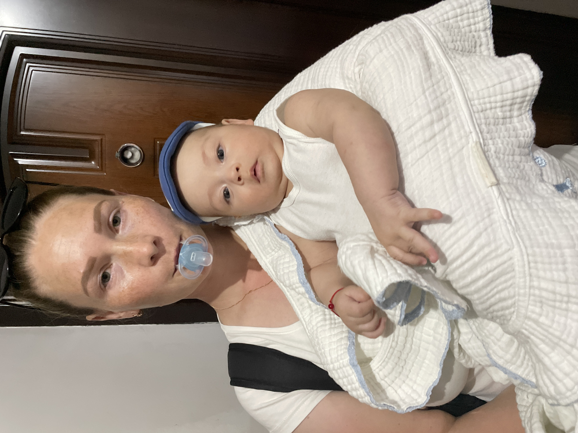 The image shows a woman holding a baby. The baby is wearing a white onesie and a blue headband. The woman is wearing a white tank top and a black strap across her chest. A blue pacifier is in the woman's mouth, and the baby is wrapped in a white blanket with blue trim. The background appears to be indoors, with a dark wooden door visible behind them. The lighting suggests it could be daytime. The overall scene is intimate, with the woman cradling the baby close to her. No visible text is present in the image.