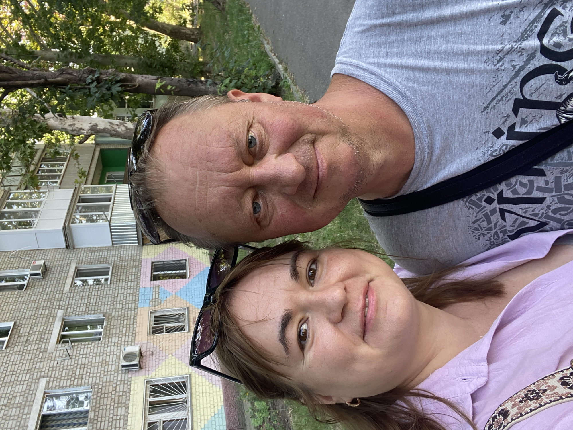 In Chornomorsk, Ukraine, a middle-aged couple takes a selfie in front of a multi-story apartment building. The building features light-colored brickwork with colorful geometric patterns in pastel shades of yellow, blue, and pink on the lower levels. Several windows, some with metal grates, are visible, along with air conditioning units mounted on the exterior. Trees with green foliage line the sidewalk, creating a backdrop of natural greenery. The man, wearing a grey t-shirt with a graphic print and sunglasses perched on his head, smiles slightly. The woman, in a light purple button-down shirt, also smiles for the camera, with her own sunglasses resting on her head. A patterned strap from a bag is visible across her shoulder. The lighting suggests it is daytime, with sunlight illuminating the scene.