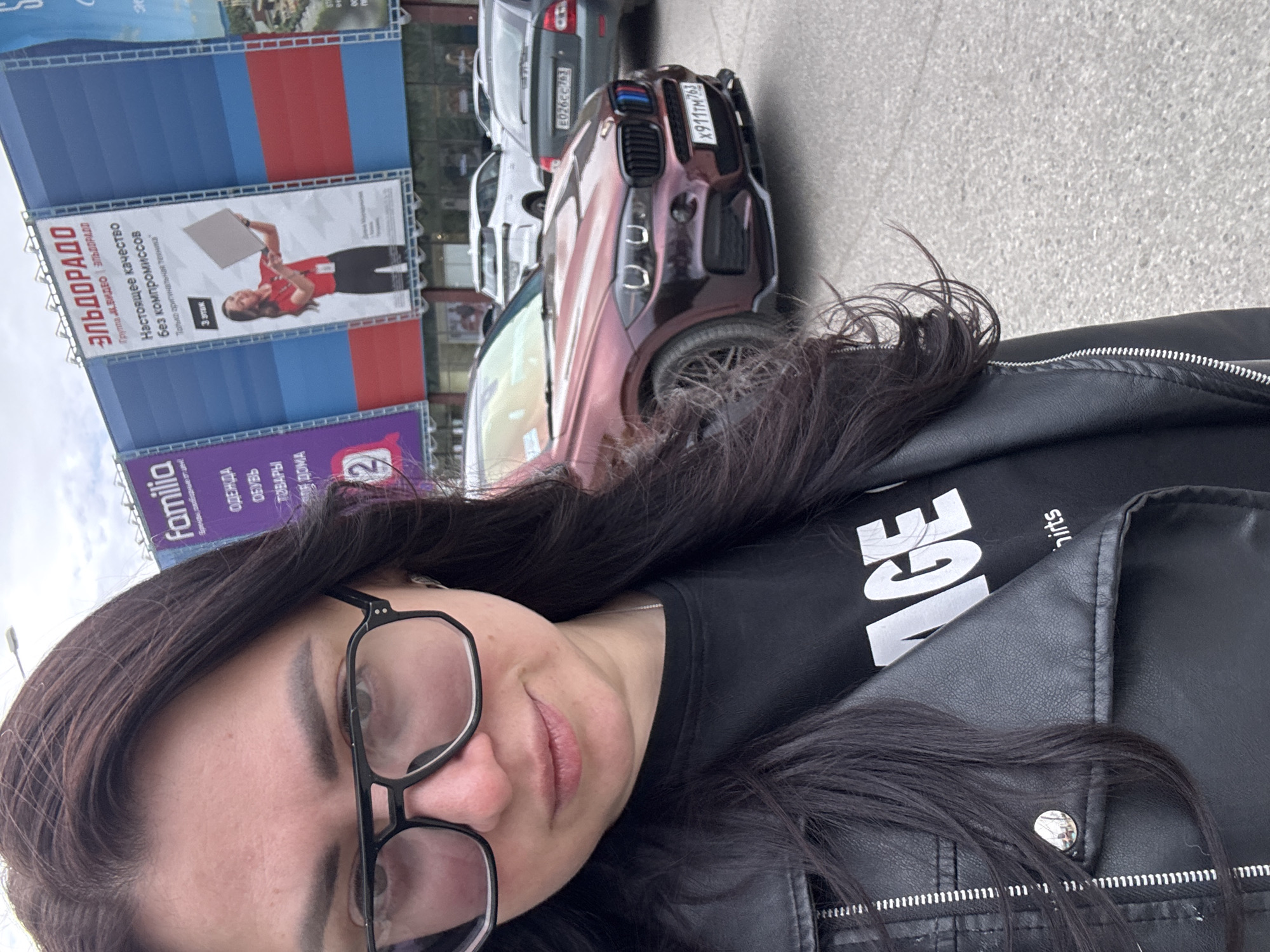 Here is a forensic description of the image:
A selfie of a woman with dark hair and glasses is at the forefront. She wears a black leather jacket over a black t-shirt. In the background, slightly out of focus, is a row of parked cars and a building with large advertisements. The building features blue and red panels with a large white advertisement for "ЭЛЬДОРАДО," featuring a woman holding a white square. Another sign below advertises "Familia," offering clothing, shoes, and home goods. A burgundy-colored car is parked close to the camera, with a partial view of its license plate "X911TM763." Another car, possibly a gray SUV, is partially visible with the license plate "E026CC763". The setting appears to be a street in an urban area during daylight.