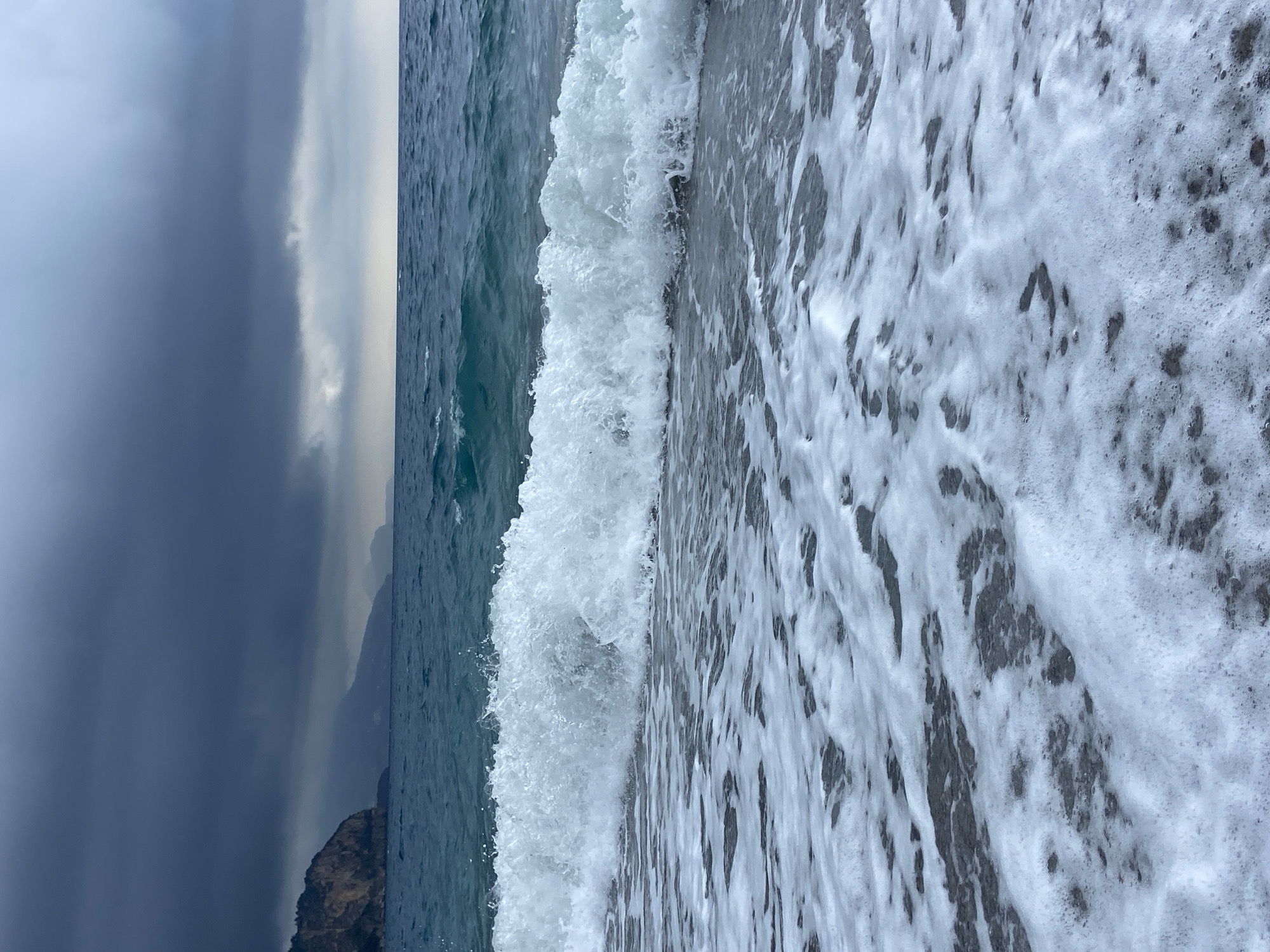 The image captures a dynamic coastal scene in Prijevor, Montenegro, under an overcast sky. The upper section of the frame is dominated by a dramatic sky, featuring heavy, dark grey clouds on the left that gradually lighten to a soft grey and white towards the right, suggesting a cloudy or stormy atmosphere.
Along the distant horizon, the faint silhouettes of mountains or cliffs are visible, blending into the dark, choppy expanse of the sea. The ocean, with its deep blue-green waters, occupies the mid-ground. A significant wave is captured in motion, actively cresting and breaking, creating a distinct line of white, frothy foam. This wave crash is a central element of the scene, illustrating the power of the sea.
The lower portion of the image depicts the immediate shoreline or beach, largely covered by a thick layer of white sea foam. Dark, wet patches, likely consisting of sand or small pebbles, are visible beneath and between the foam, adding texture to the foreground. The overall impression is one of a rugged, natural coastline on a dramatic, cloudy day. There are no people or visible text in the image.