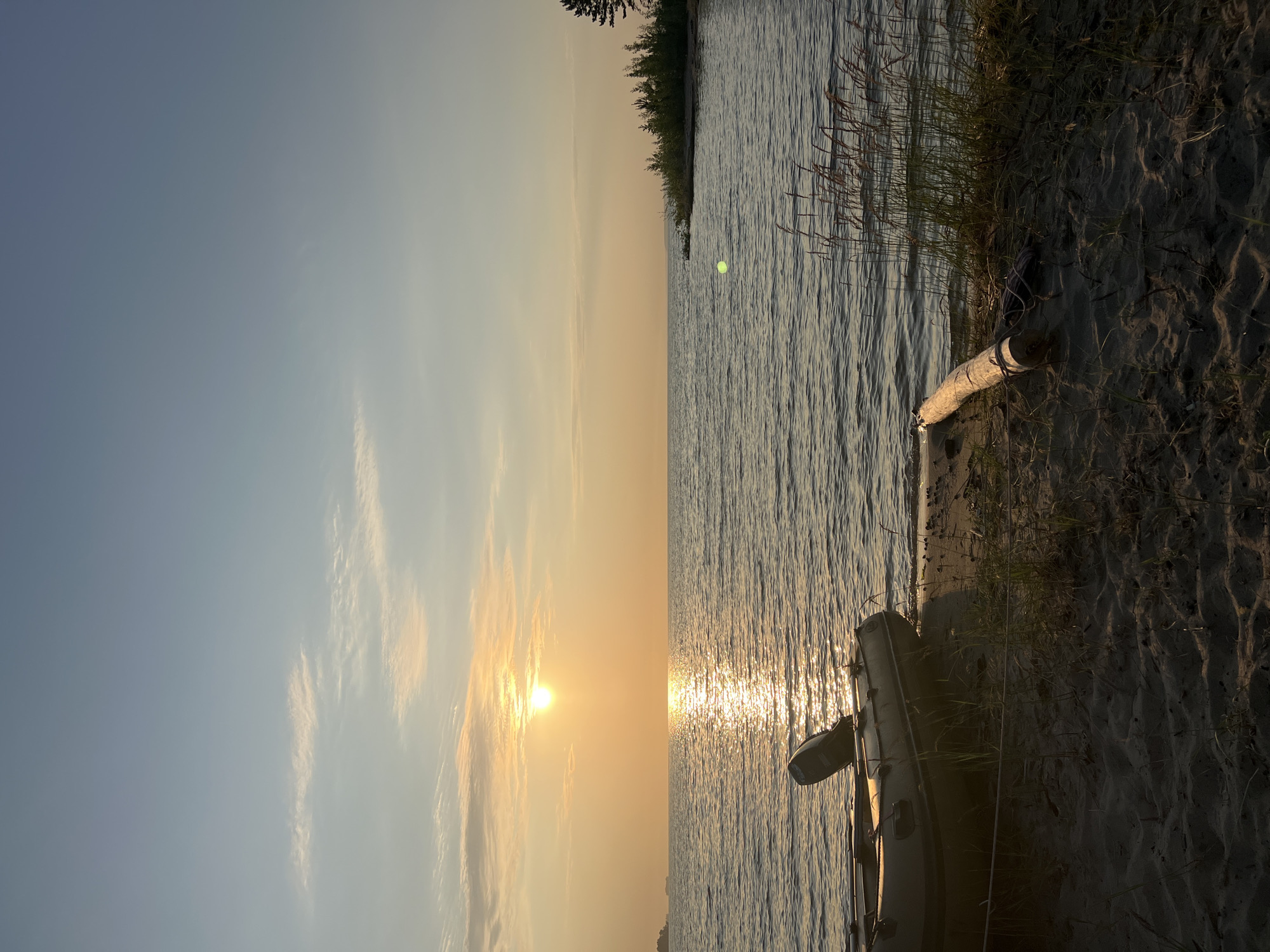 An inflatable boat with an outboard motor rests on a sandy shore by a body of water. The sun is setting, casting a warm glow across the sky and reflecting on the water's surface. The scene appears to be located near the city of Borovoye, Russia, with a distant shoreline visible in the background. There are no people present.