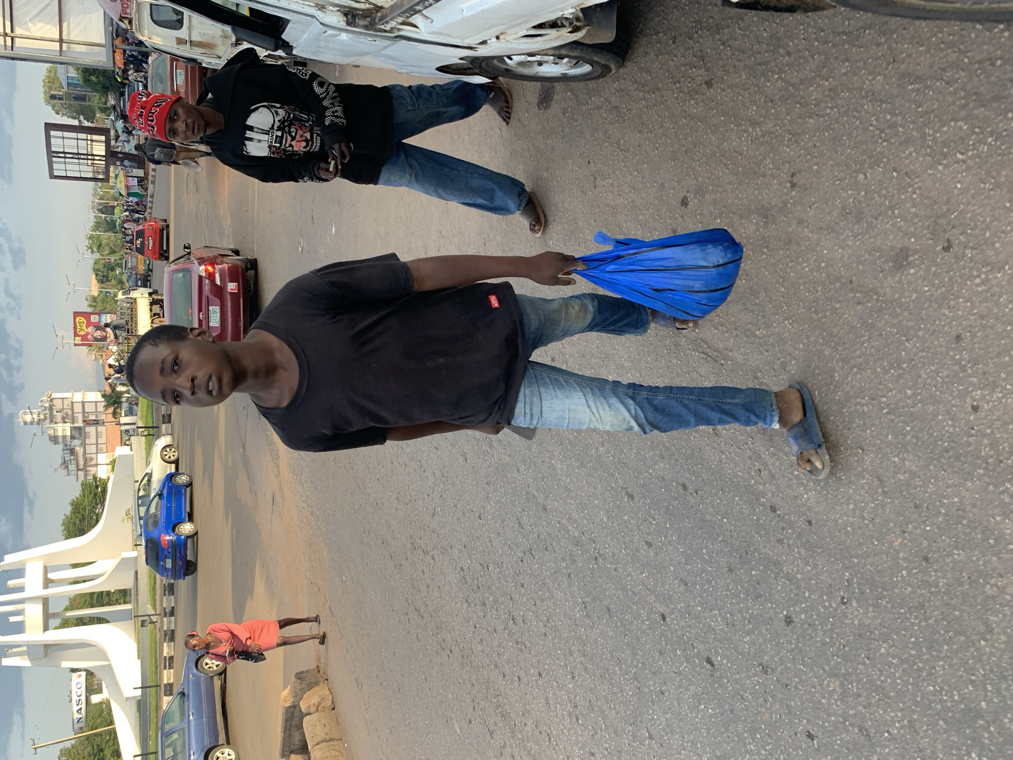 The image shows a street scene in Jos, Nigeria. Two young men are the primary subjects. One is wearing a black hoodie with graphics and a red beanie, and is standing next to a white vehicle. He is holding a cigarette and looking towards the camera. The second young man, in the foreground, is wearing a black t-shirt and blue jeans. He is holding a folded blue object, possibly an umbrella or a bag, and is also looking at the camera.
In the background, several vehicles are visible, including a red car and a blue car. A large white monument, potentially a landmark, is situated on the left side of the image. The overall setting appears to be a busy urban area, possibly a market or a transport hub, with a considerable amount of traffic and pedestrian activity suggested by the presence of multiple cars and people in the distance. A woman in an orange dress is also visible walking near a blue car.
The time of day appears to be late afternoon or early evening, indicated by the soft lighting and long shadows. The weather seems to be clear, with a visible sky in the upper portion of the image.
One visible text element is "NASCO" printed on a white sign behind the monument. The overall mood is candid, with the subjects acknowledging the camera. The scene depicts everyday life in an urban environment.