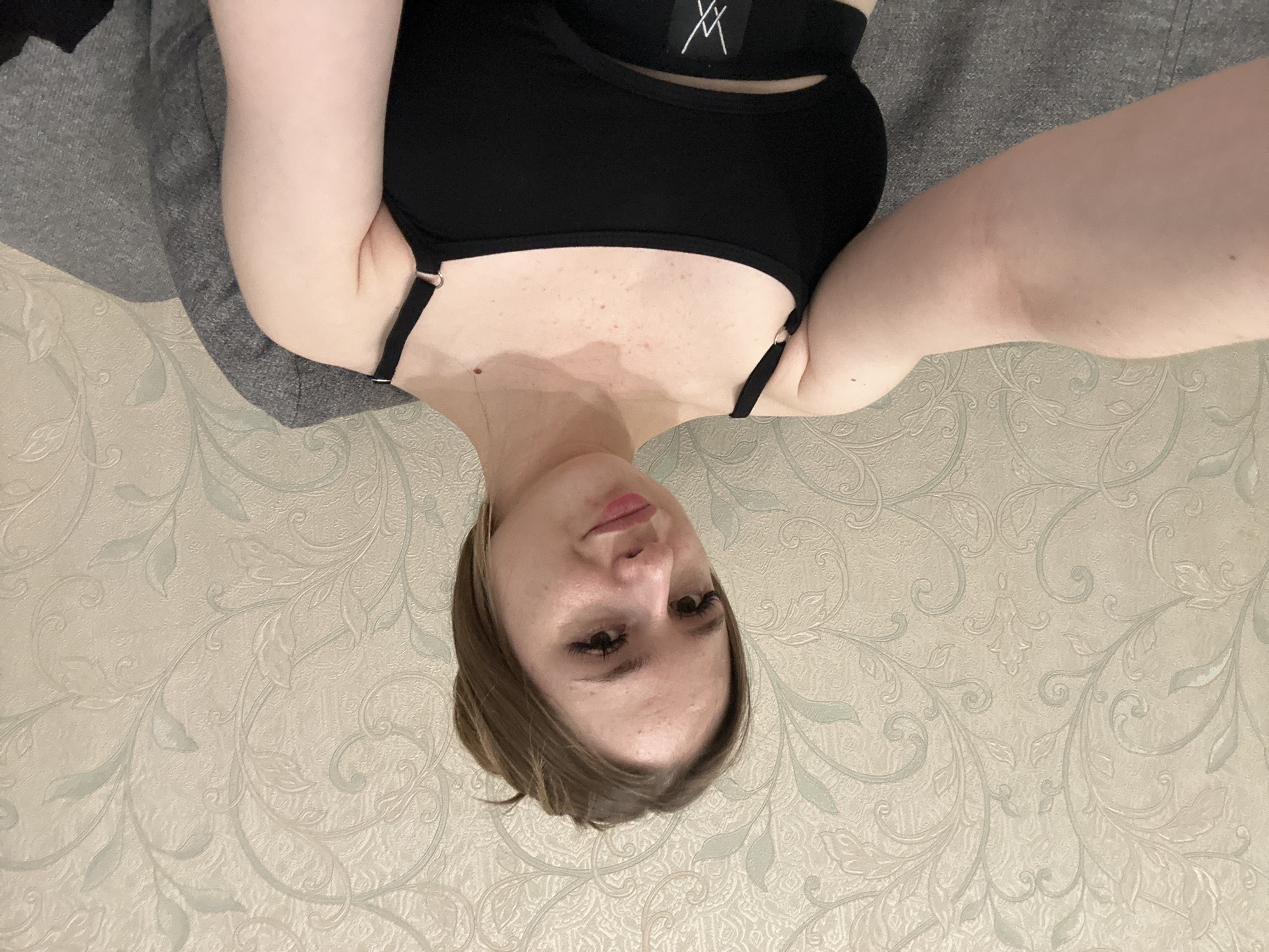 A young woman with light brown hair pulled back is taking a selfie in a dimly lit room. She is wearing a black tank top and sitting on a gray couch in front of patterned wallpaper. Her left arm is extended towards the camera, and her right arm is resting on the couch. She has makeup on, including eyeliner and mascara, and is smiling slightly at the camera.