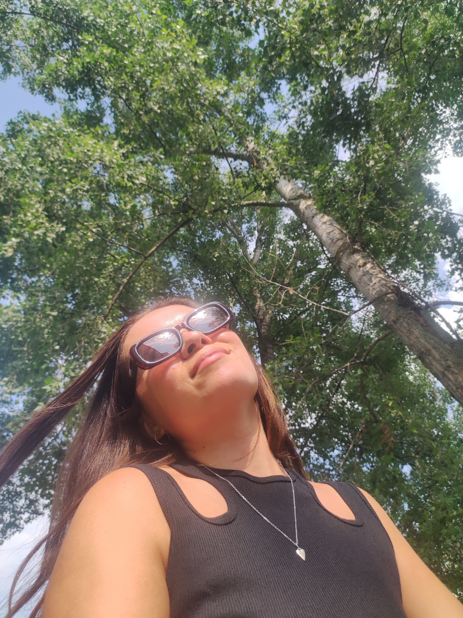 The image shows a young woman from the shoulders up, looking upwards. She is wearing sunglasses and a black tank top with a cut-out detail at the neckline, and a silver pendant necklace. Her long brown hair is blowing to the left. The background is filled with the dense green leaves and branches of a tree, with glimpses of a blue sky. The perspective is from below, looking up into the canopy. The scene appears to be outdoors in a natural setting, possibly a park or wooded area within the city of Chaikovsky, Russia.