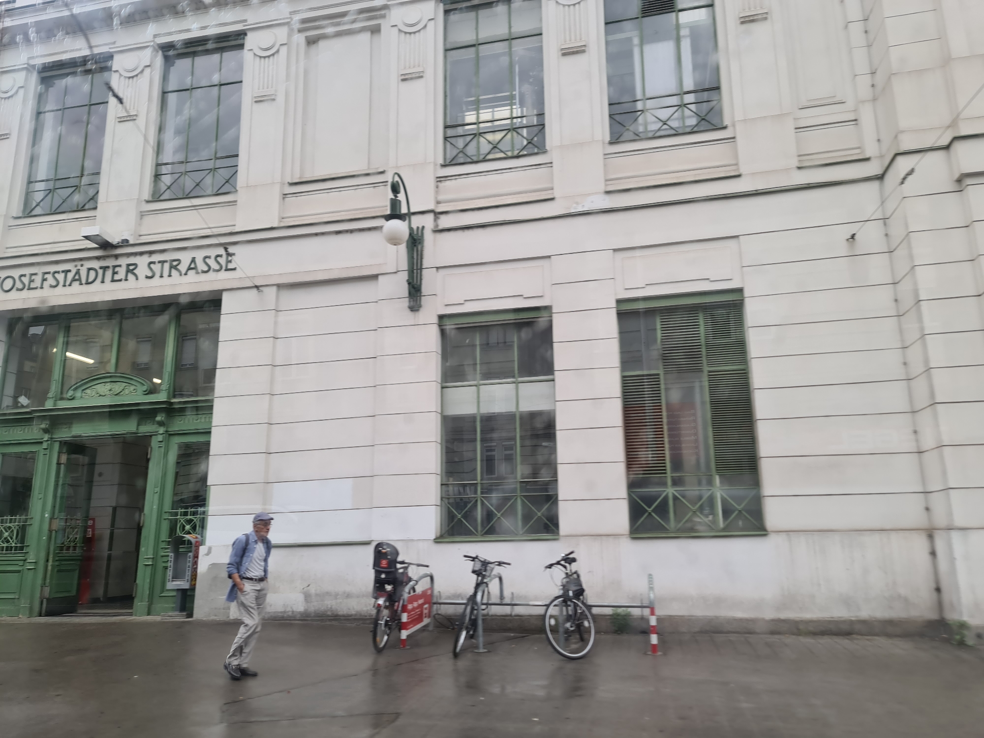 The image shows a street scene in Vienna, Austria, likely in front of a building with the inscription "JOSEFSTÄDTER STRASSE". An elderly man, wearing a blue shirt, beige pants, and a blue cap, is walking on the wet sidewalk. To his right, there is a row of bicycles parked at a bike rack. The building is modern with large windows. The entrance to a building on the left is adorned with green decorative elements and glass doors. The overall scene appears to be captured during the daytime, with overcast weather conditions.