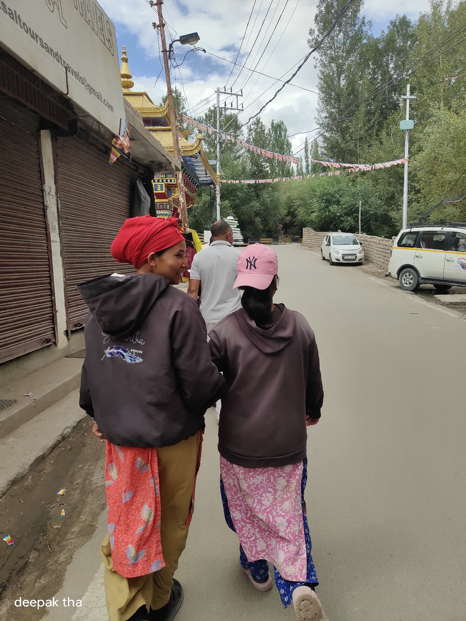 In Leh, India, two women walk away from the camera down a paved street. The woman in the foreground wears a red turban, a black hooded jacket with a blue feather design on the back, and a red patterned cloth wrapped around her waist over khaki pants. The woman behind her wears a pink baseball cap with a black "NY" logo, a brown hooded jacket, and a pink and white floral patterned garment over a blue patterned fabric. A man in a grey t-shirt walks ahead of them, also moving away from the camera. To the left of the women are closed metal shutters of a shop with a sign displaying text including "sal tour sand travels@gmail.com". Further down the street, a white car is parked and a white van with yellow trim is visible. Prayer flags are strung across the street and on the roofs of buildings decorated with golden stupas. The environment appears to be an urban street with trees lining the background under a cloudy sky.