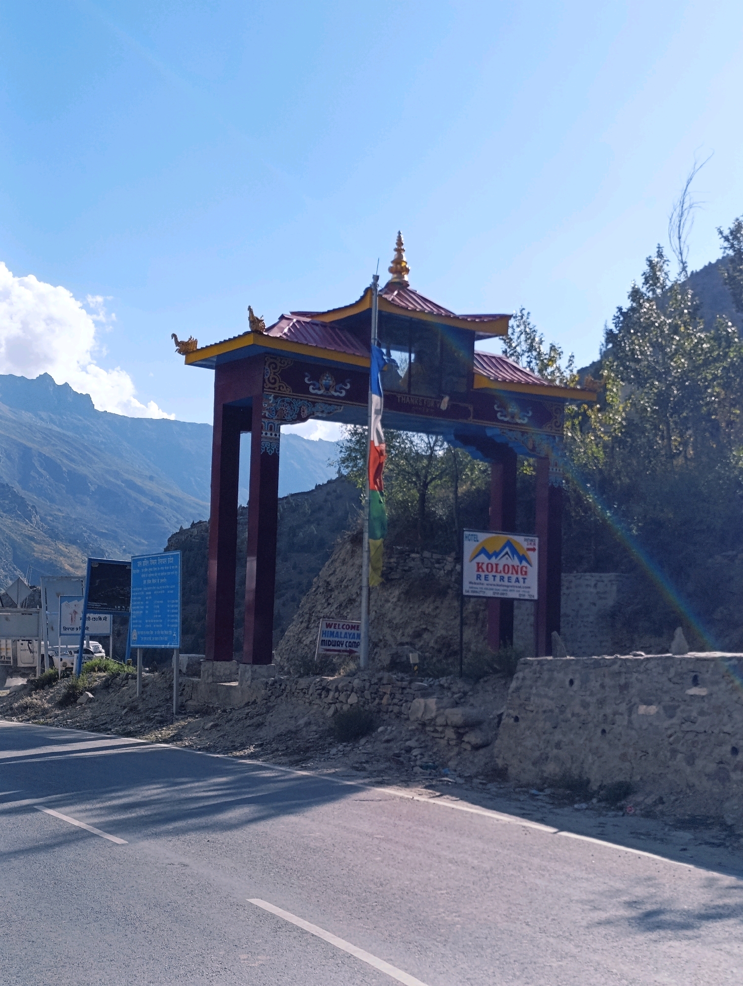 The image captures an ornate gateway in Kead, India, situated against a backdrop of majestic mountains and a clear blue sky. The gateway, painted in vibrant red and adorned with golden detailing, features traditional Buddhist architectural elements. A colorful flag with multiple stripes, possibly representing Buddhist prayer flags, is seen fluttering from the gateway's structure.
To the left of the gateway, several informational signs are mounted on poles. One prominent sign, predominantly blue, displays text in Hindi, likely conveying local information or directions. Another sign, black and white, also presents text in Hindi.
To the right of the gateway, a sign advertises "KOLONG RETREAT HOTEL," featuring a mountain and sun graphic. Below this, more text provides contact details and potentially a website address. Further to the right, another sign reads "WELCOME HIMALAYAN MIDWAY CAMP."
The foreground shows a paved road with distinct white lane markings, suggesting a highway or major route. Shadows of trees are cast across the road, indicating sunlight filtering through the foliage of trees lining the right side of the scene. The overall environment suggests a scenic, possibly mountainous region, typical of Northern India.