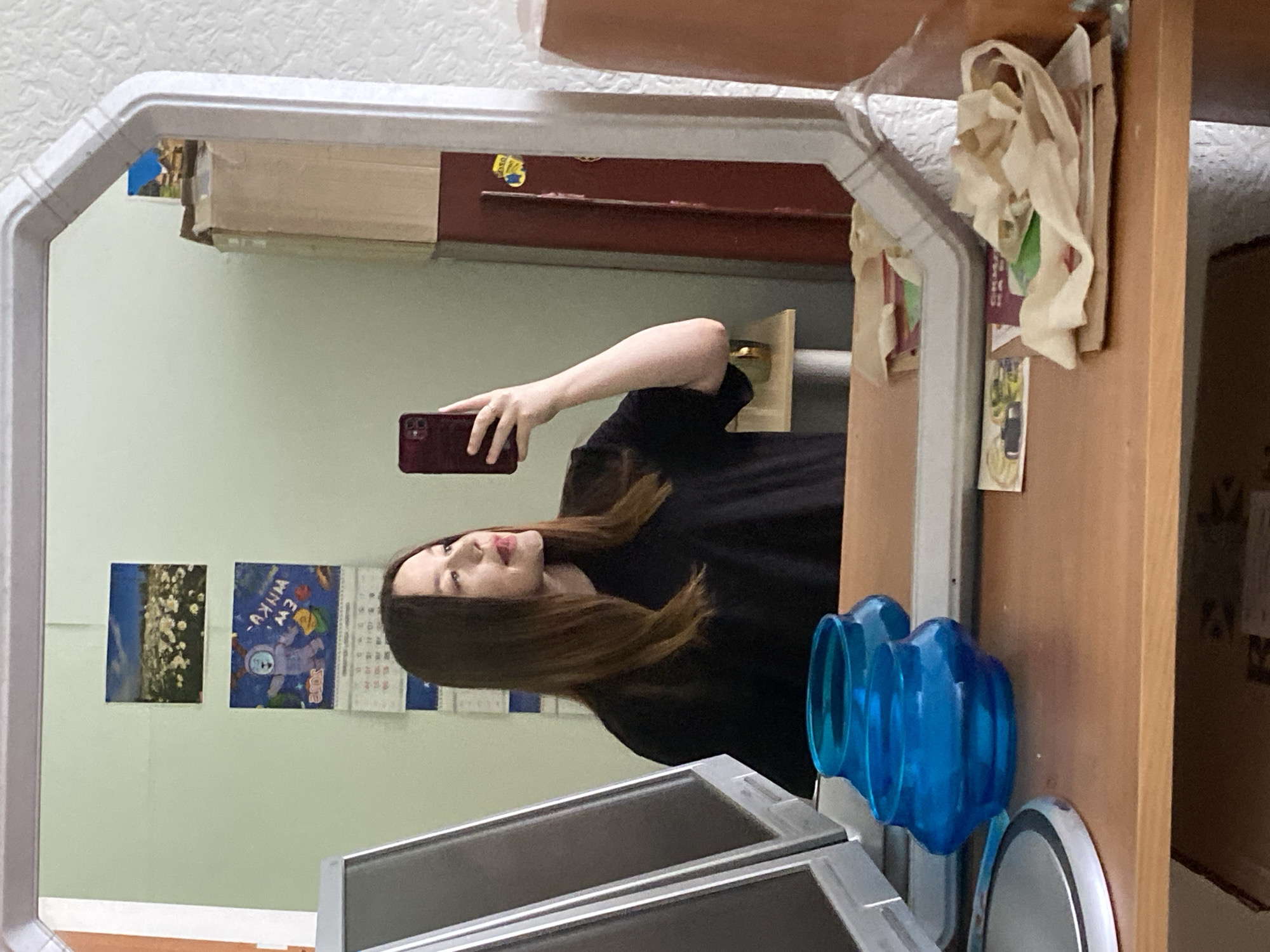 A person is taking a selfie in a mirror. The mirror reflects the individual holding a phone, a wall with pictures and a calendar, and part of a room. Items are on a table next to the mirror, including blue plastic objects and a white container. The location is Chaikovsky, Russia, based on the 2018 calendar reflected in the mirror and the Cyrillic script on the calendar.