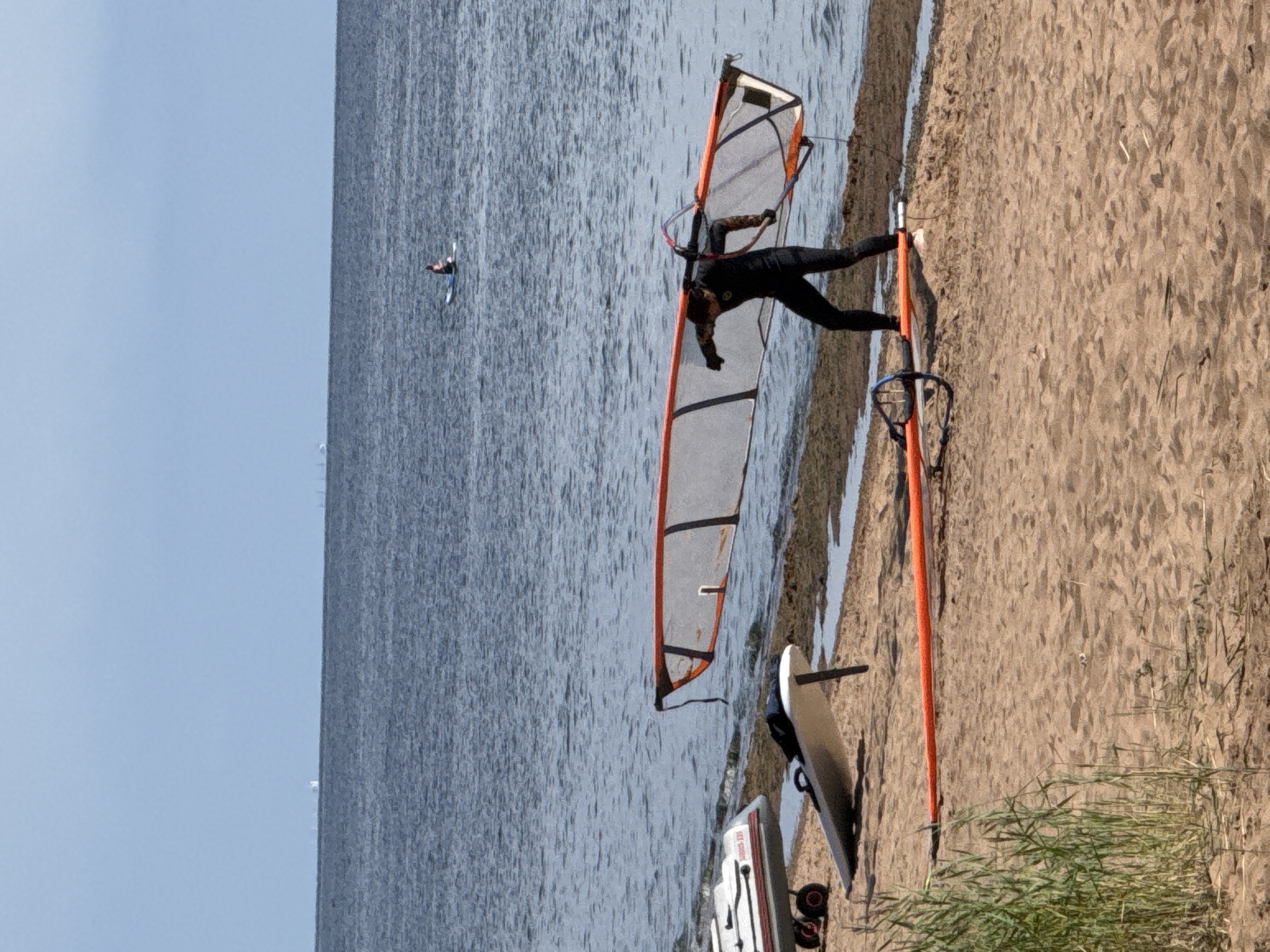 In the city of Zelenaya Roshcha, Russia, a person wearing a black wetsuit stands on a sandy beach, carrying a windsurfing sail over their shoulders. The sail is mostly white with orange trim. To the left of the person, two windsurf boards are visible on the sand. One board is white and lies on its side with its fin sticking up. Another larger object, possibly a boat or raft on wheels, is partially visible to the far left. In the background, the calm, grey-blue water of a large body of water stretches to the horizon under a pale blue sky. A single individual is seen on a paddleboard in the distance. The overall scene depicts a calm day at the beach with equipment prepared for windsurfing.