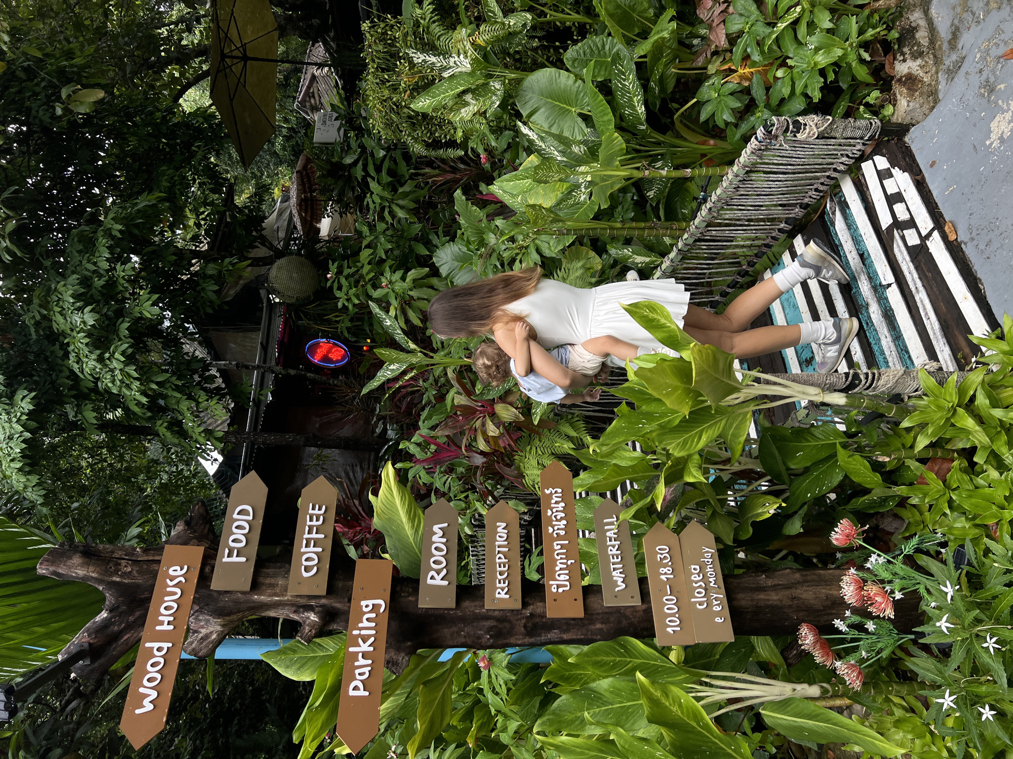 A woman in a white dress carries a young child while walking on a wooden bridge in Choeng Thale, Thailand. The bridge has white and blue painted planks and rope railings. To the left of the bridge is a wooden signpost with multiple directional arrows. These arrows point to "Wood House," "FOOD," "COFFEE," "Parking," "ROOM," "RECEPTION," Thai script, "WATERFALL," and operational hours. The signpost is surrounded by lush green tropical foliage. In the background, there are buildings, umbrellas, and more dense vegetation.