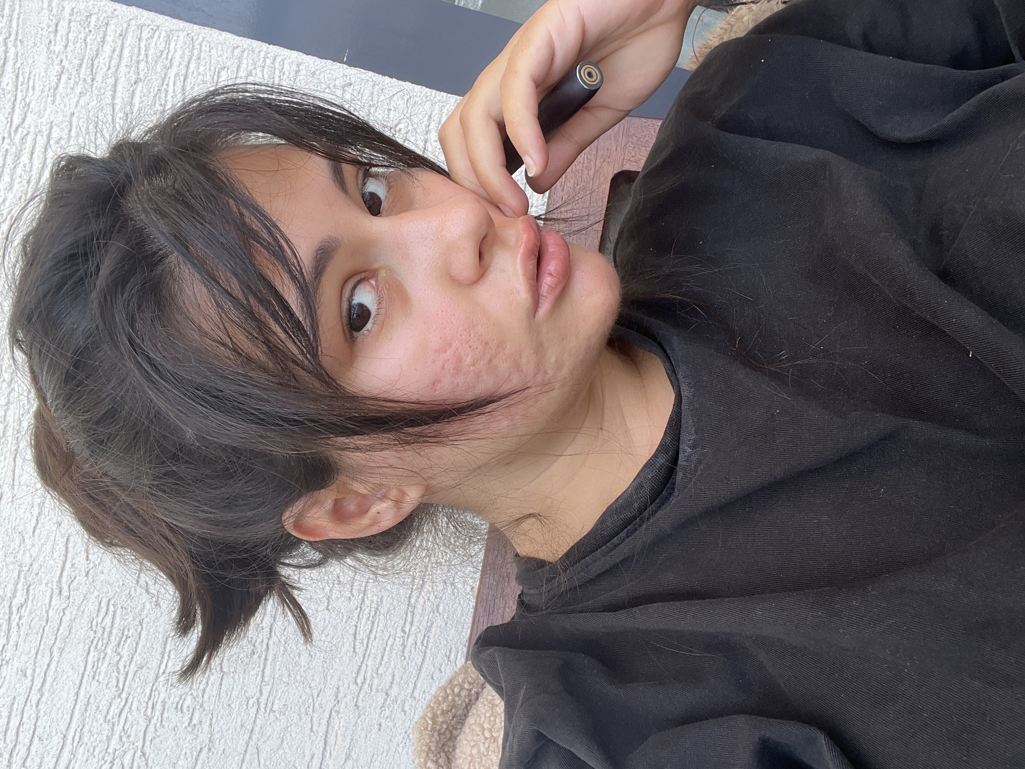 A young woman with dark hair pulled back into a messy bun, bangs falling across her forehead, is captured in a close-up selfie. She is wearing a black sweatshirt. Her left hand rests near her mouth, holding a dark cylindrical object with a metallic tip. Her skin, particularly on her left cheek, shows signs of acne. The background features a textured white wall and a hint of dark material, suggesting an outdoor or semi-outdoor setting.
