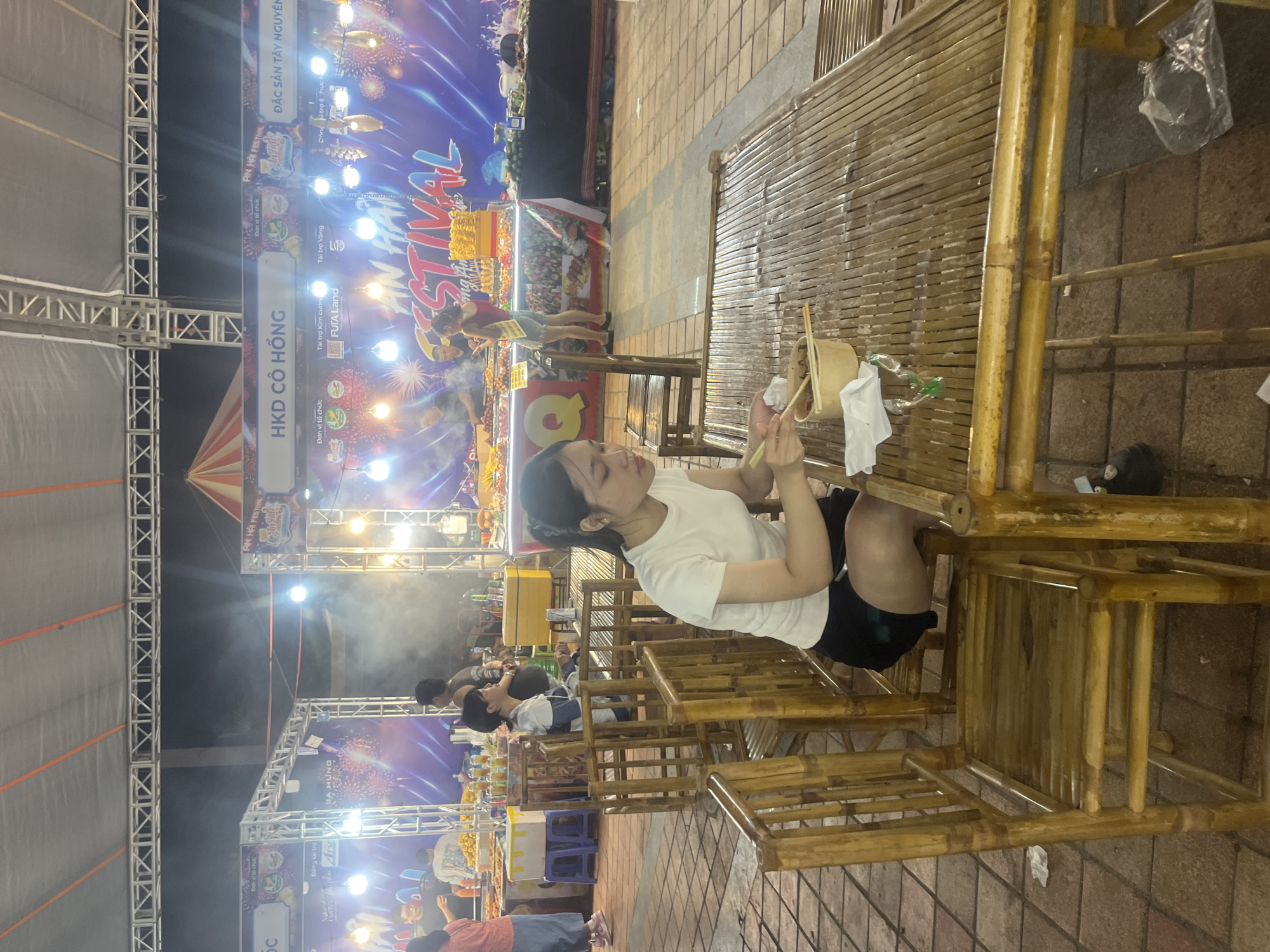 In Đà Nẵng, Vietnam, a young woman with dark hair pulled back is seated at a bamboo table, actively eating from a bowl with chopsticks. She is wearing a white t-shirt and black shorts. The setting appears to be an outdoor night market or festival, as evidenced by the strung lights, food stalls in the background with vibrant banners and displays of food, and people milling about. One banner displays "AN HAI FESTIVAL". The ground is paved with stone tiles. A few empty bamboo chairs are visible around the table. Smoke emanates from one of the food stalls in the background, suggesting cooking is taking place.