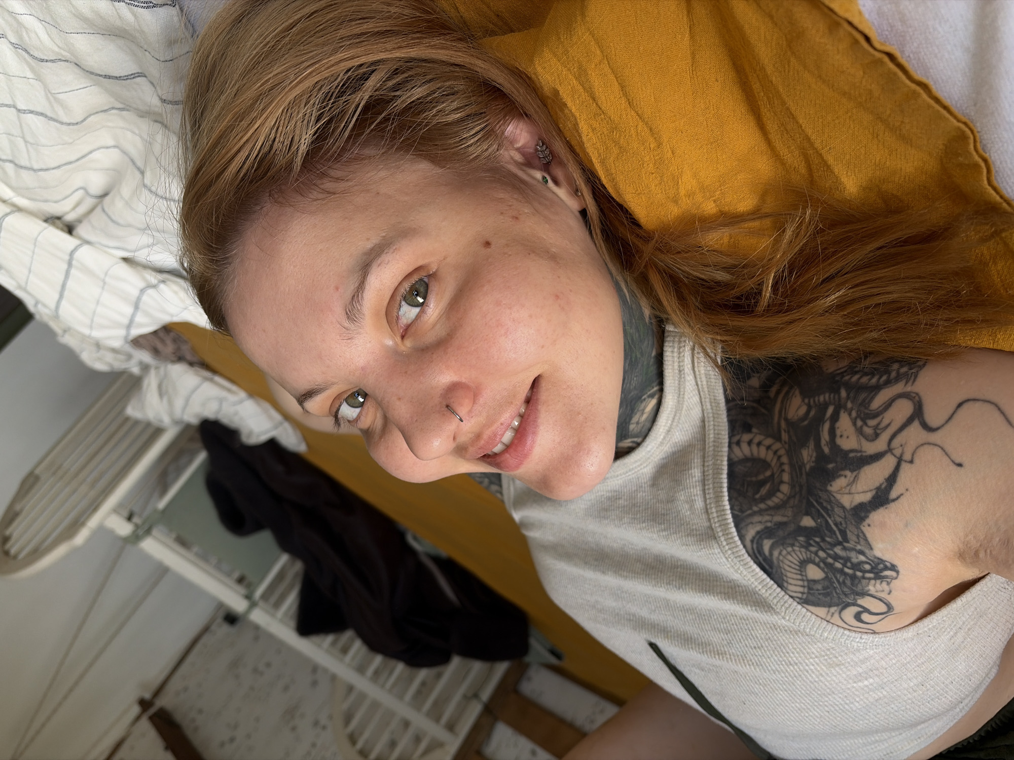 A blonde-haired person lies on a bed in what appears to be an interior setting. The person is smiling slightly, with a nose ring and multiple ear piercings visible. A large black tattoo is visible on the person's chest and neck. The bed is covered with striped bedding and a yellow pillow. A cot with a black shirt is visible in the background. The location is in a city called Chaikovsky, Russia.