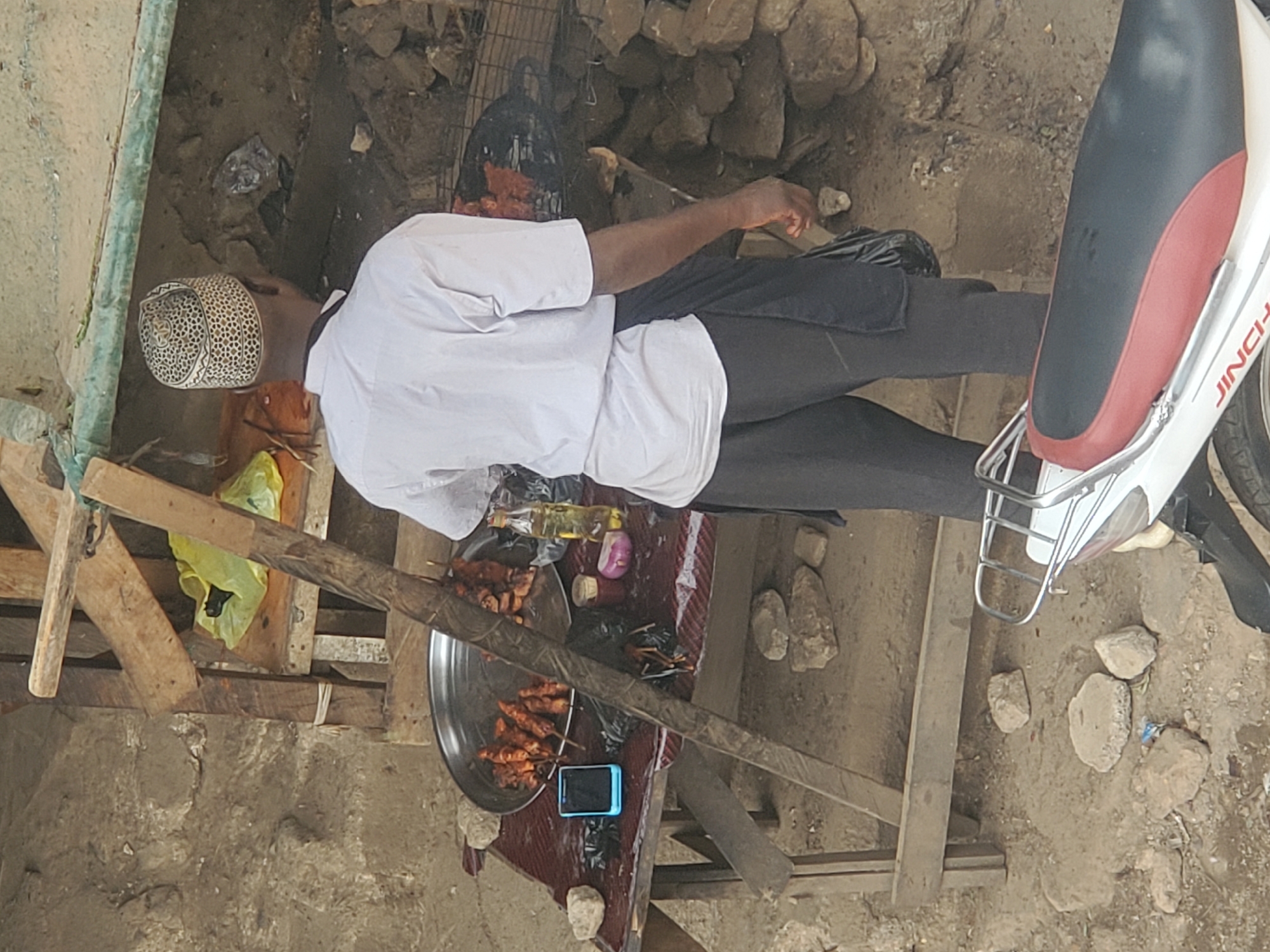 A man wearing a white shirt and patterned kufi is grilling meat skewers on a small grill. The grill is placed on a bed of rocks and debris. Next to him, skewers of grilled meat are displayed on a round tray on a makeshift wooden table. A cell phone is on the table near the food. The scene is on a dirt ground with building materials and debris scattered around. The back of a motorcycle is visible to the right of the man. This is likely a street-side food vendor scene in Jos, Nigeria.