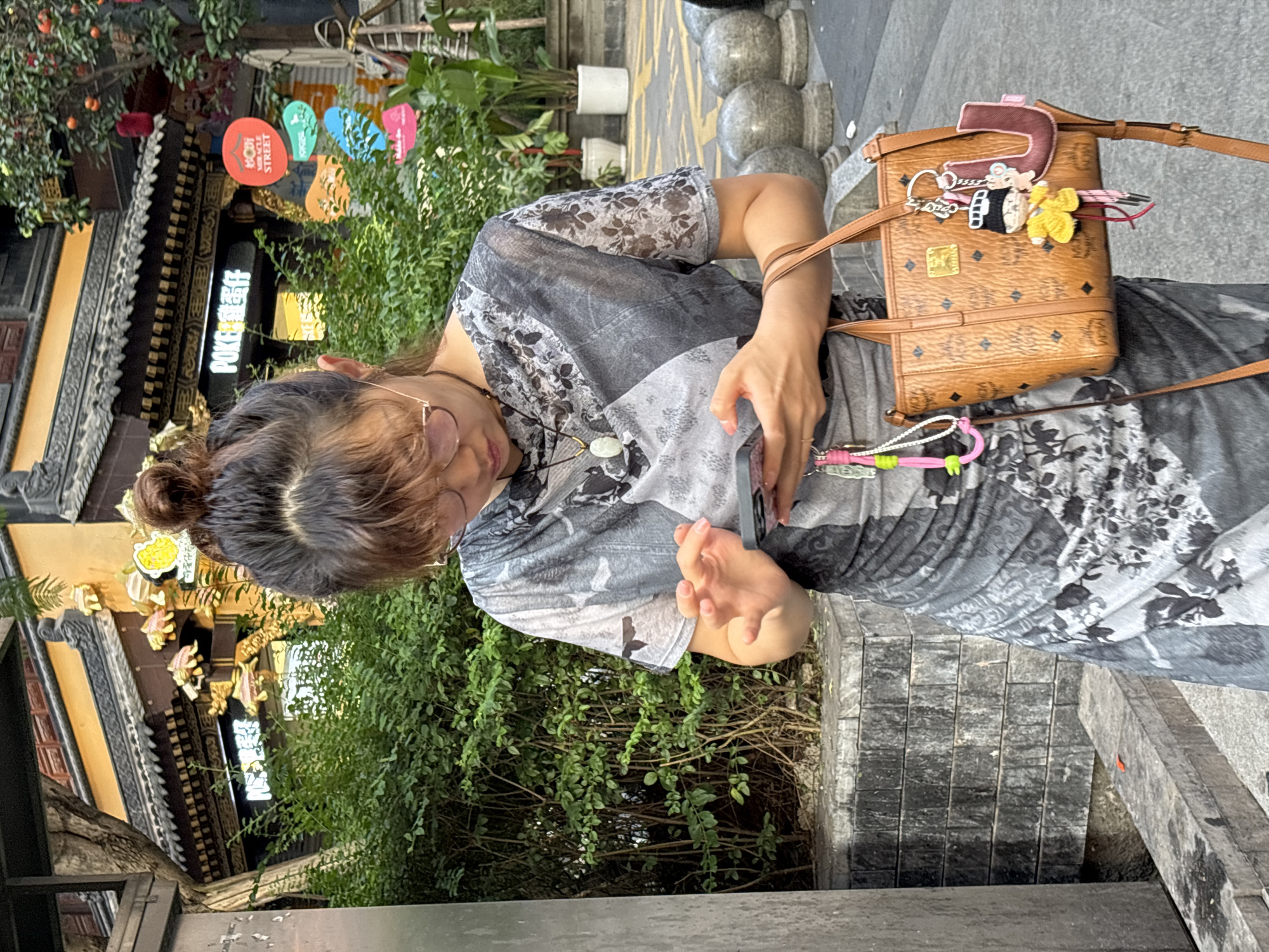 A woman stands on a street in Yuzhong District, China, looking down at her phone. She is wearing a grey and black patterned dress and glasses. She has a brown MCM handbag with several charms attached. The background shows traditional Chinese architecture with a tree and lush greenery.
