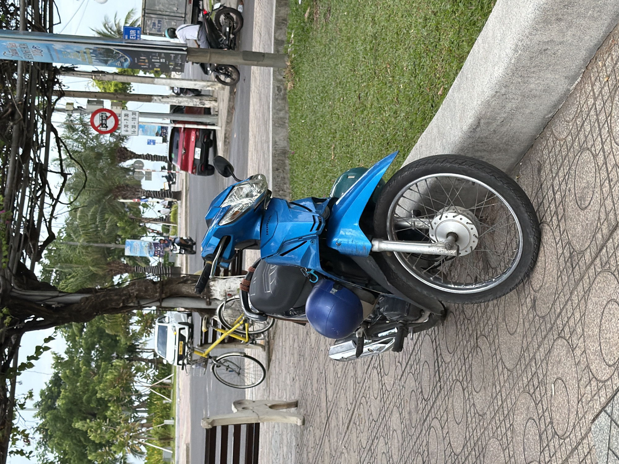 The image depicts a street scene in Nha Trang, Vietnam, likely near a coastal area given the presence of palm trees and vegetation. In the foreground, a bright blue motorcycle is parked on a tiled sidewalk. A blue helmet is attached to the side of the motorcycle. Behind the motorcycle, a yellow bicycle with a basket is leaning against a tree. Further back, a white car is visible on the road, along with other vehicles including a red SUV and a truck. Traffic signs are present, indicating traffic regulations. People are seen walking in the background and riding motorcycles. The environment is characterized by trees, green grass, and a clear sky, suggesting it is daytime. The architecture and signage also point towards a Southeast Asian urban setting.