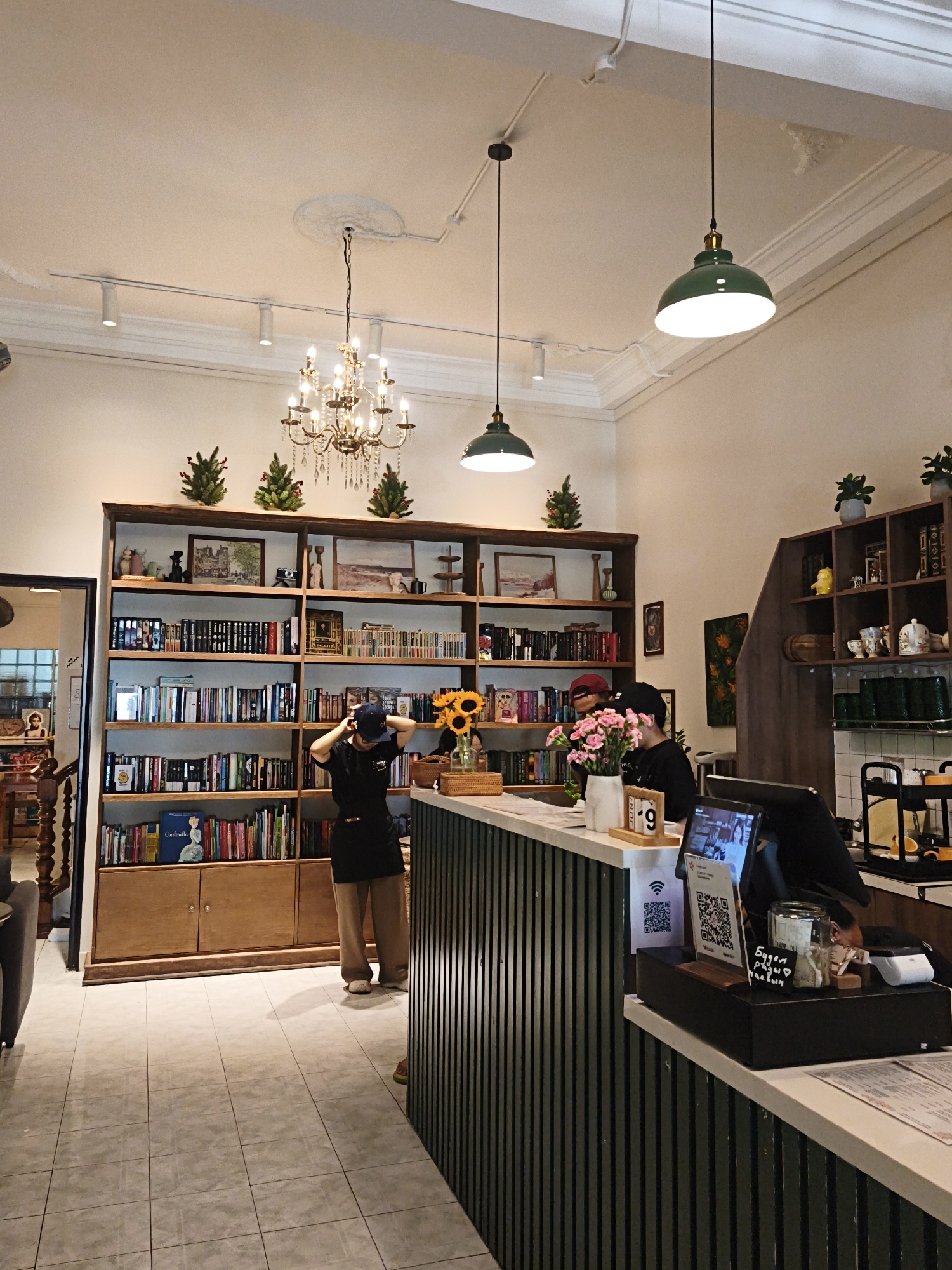 This image depicts the interior of a cafe in Bac Nha Trang Ward, Vietnam. The cafe features a warm and inviting atmosphere, with light-colored walls and a tiled floor. A large wooden bookshelf filled with books and decorative items dominates the left side of the frame, showcasing a collection of novels, art books, and trinkets. A classic chandelier hangs from the ceiling, casting a soft glow. Two pendant lights with green shades are also visible.
In the foreground, a counter with a white top and a dark, ribbed base serves as the central point of activity. Two individuals are behind the counter, appearing to be staff members. One person, wearing a black t-shirt and khaki pants, is adjusting their hat. The other person, also in dark attire, is partially obscured by the counter. In front of the counter, a tablet displays an image, and a small wooden calendar shows "JUNE 9". A vase of pink flowers and a basket of sunflowers add a touch of floral decor. A stack of menus rests on the counter.
The background reveals more shelves filled with various items, including teapots, cups, and other decorative objects. A large painting with an abstract floral design hangs on the wall. The overall impression is that of a cozy and well-decorated cafe, likely a popular spot for locals and tourists alike.