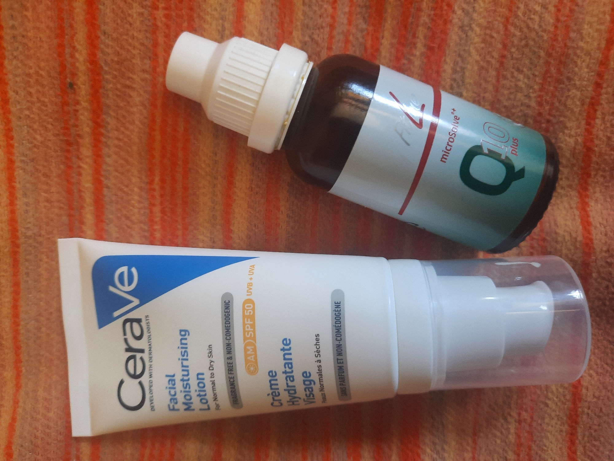 Two cosmetic products, a tube of CeraVe Facial Moisturising Lotion and a small bottle of what appears to be a Q10 supplement, are placed diagonally on a textured surface with orange and cream stripes. The CeraVe tube is white with blue and black lettering, and it has a pump dispenser. The supplement bottle is dark glass with a white cap and a label that includes the text "Q10 plus". The image is shot from a high angle, and there are no people or other objects visible. The environment is indeterminate.