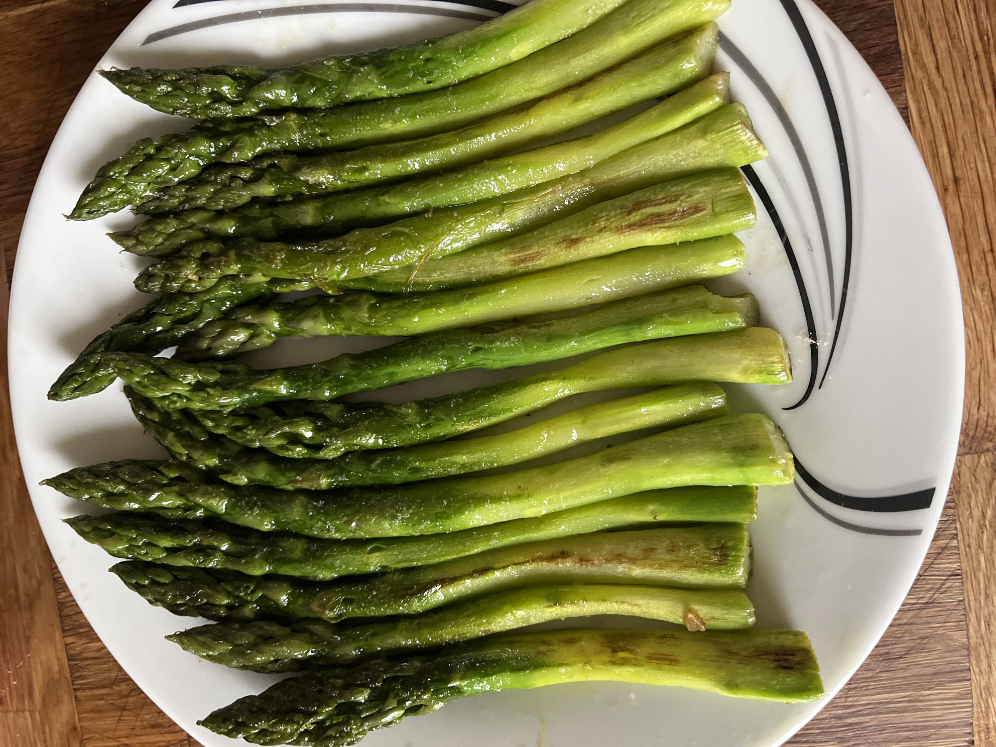 The image presents an overhead view of a serving of cooked green asparagus spears neatly arranged on a white plate. The asparagus exhibits a vibrant green color and appears glistening, likely from oil or moisture. Small white crystalline specks, consistent with salt, are visible on many of the stalks, indicating seasoning. Some spears, particularly towards their thicker ends, show light brown or reddish-brown marks, suggesting they might have been roasted, grilled, or pan-fried. The delicate tips of the asparagus are visible on the left side of the plate, while the trimmed ends point towards the right.
The white ceramic plate features a distinctive design on its right side, consisting of several curved, parallel stripes in varying shades of black and grey. A warm-toned wooden surface, possibly a table or cutting board, forms the background, visible in the upper and lower right corners of the frame. The lighting is bright and even, illuminating the food and enhancing its textures. There are no people, explicit activities, or visible text in the image. The overall scene conveys a sense of a freshly prepared, healthy dish, ready for consumption.