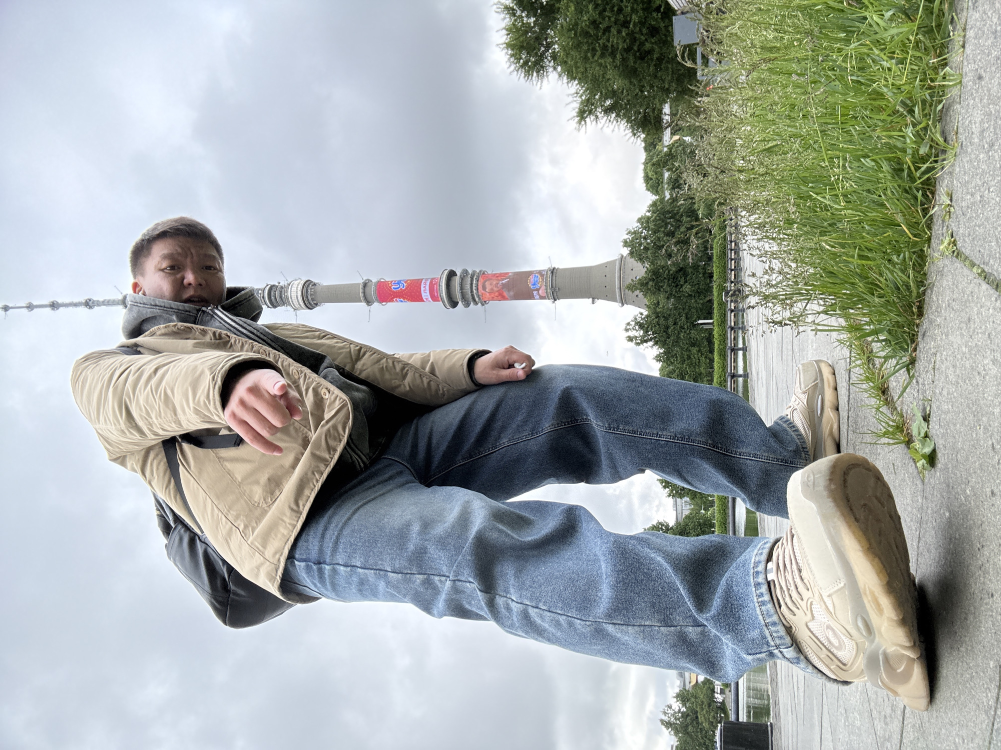 A low-angle shot depicts a person with short brown hair wearing a beige jacket, blue jeans, and beige sneakers. The person is positioned as if reclining or leaning back, with one foot extended towards the viewer. The Ostankino Tower is visible in the background, decorated with advertisements. A concrete walkway and patches of greenery are present. The sky is overcast. The location is in Khimki, Russia.