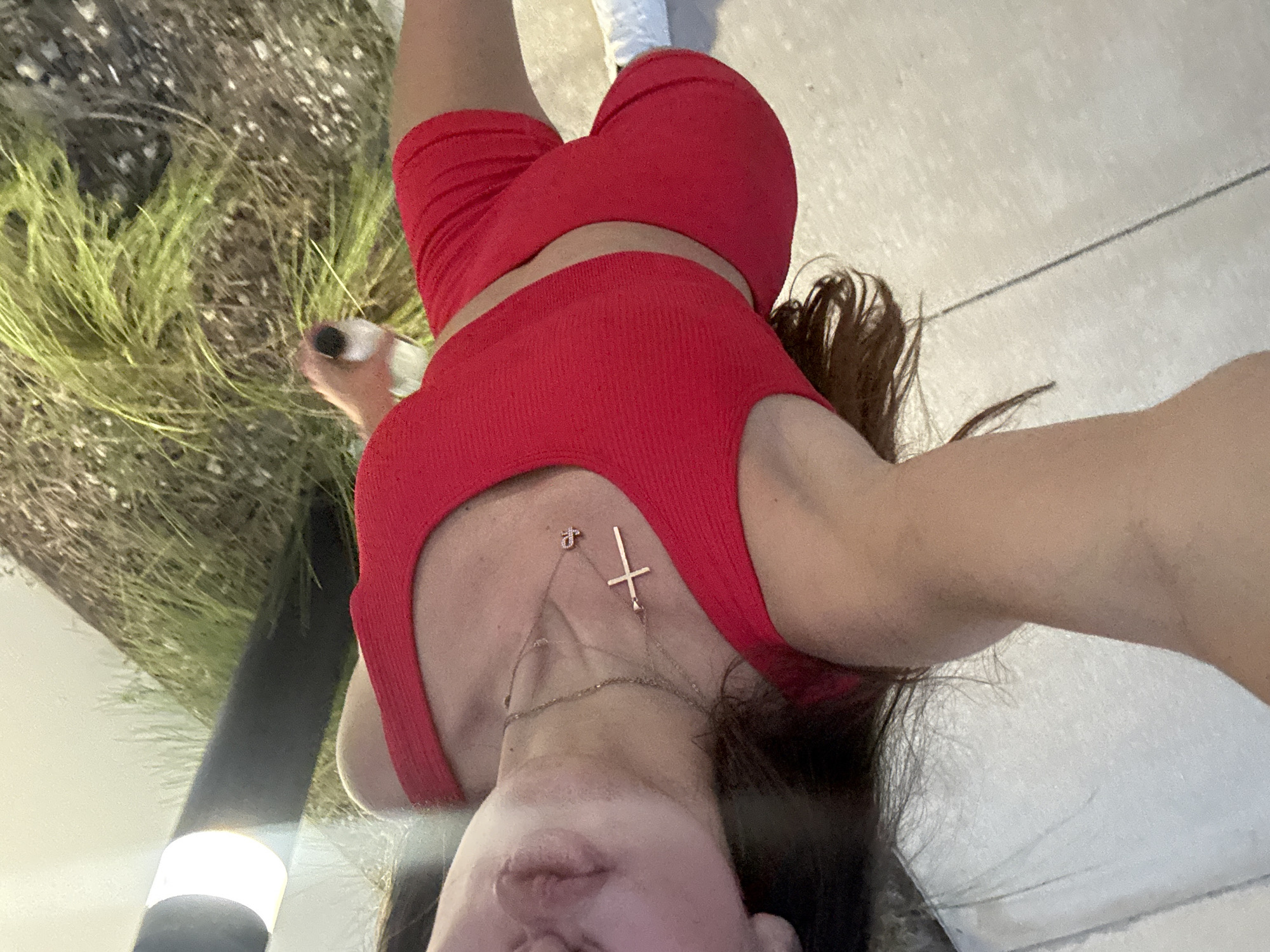 A person, viewed from the shoulders down, is wearing a red athletic set, consisting of a crop top and shorts, and white sneakers. They are holding a clear bottle with a black lid in their right hand. The person is standing on a tiled surface, with a patch of tall grass and a black pole with a light fixture in the background. The image is oriented vertically, with the person's head at the top left of the frame.
