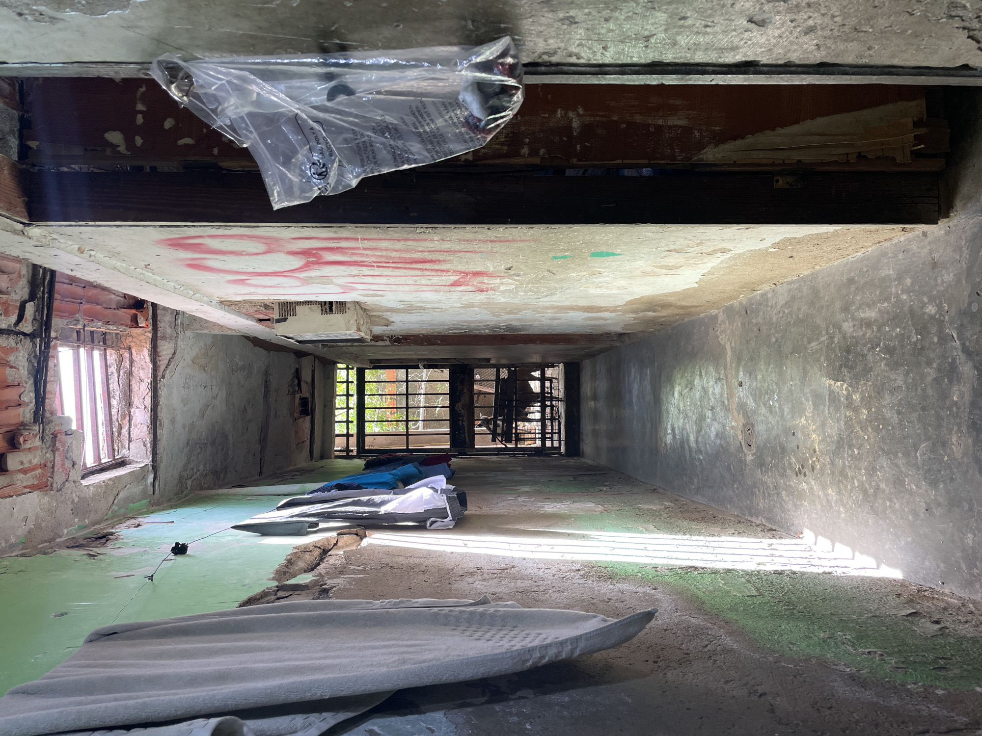 A low-angle shot looking down a dilapidated hallway in what appears to be an abandoned or severely neglected building. The hallway is narrow, with bare, crumbling walls and a partially deteriorated floor. On the left, brick walls are visible, interspersed with broken window frames. Graffiti is visible on the ceiling above. At the end of the hallway is a dark, barred opening leading to the outside, where greenery is vaguely visible. Several items of bedding, including sheets and blankets, are scattered along the floor of the hallway. There are no people visible. The location appears to be within a building or structure; no specific city is identified.
