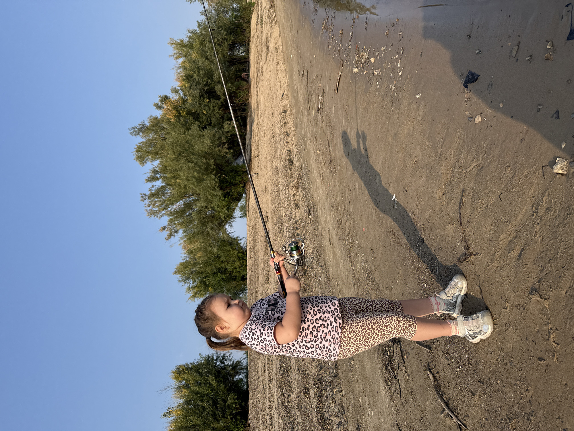 A young Caucasian girl with brown hair pulled into a ponytail is standing on a sandy embankment, holding a fishing rod. She is wearing a pink t-shirt with a leopard print, leopard print leggings, and grey and white sneakers with pink trim. The fishing rod is extended horizontally, and she is holding it with both hands. The background consists of a sandy slope with sparse vegetation and trees. The sky is clear and blue. The location is the bank of a body of water, likely a river or lake. The image captures a moment of outdoor recreation.