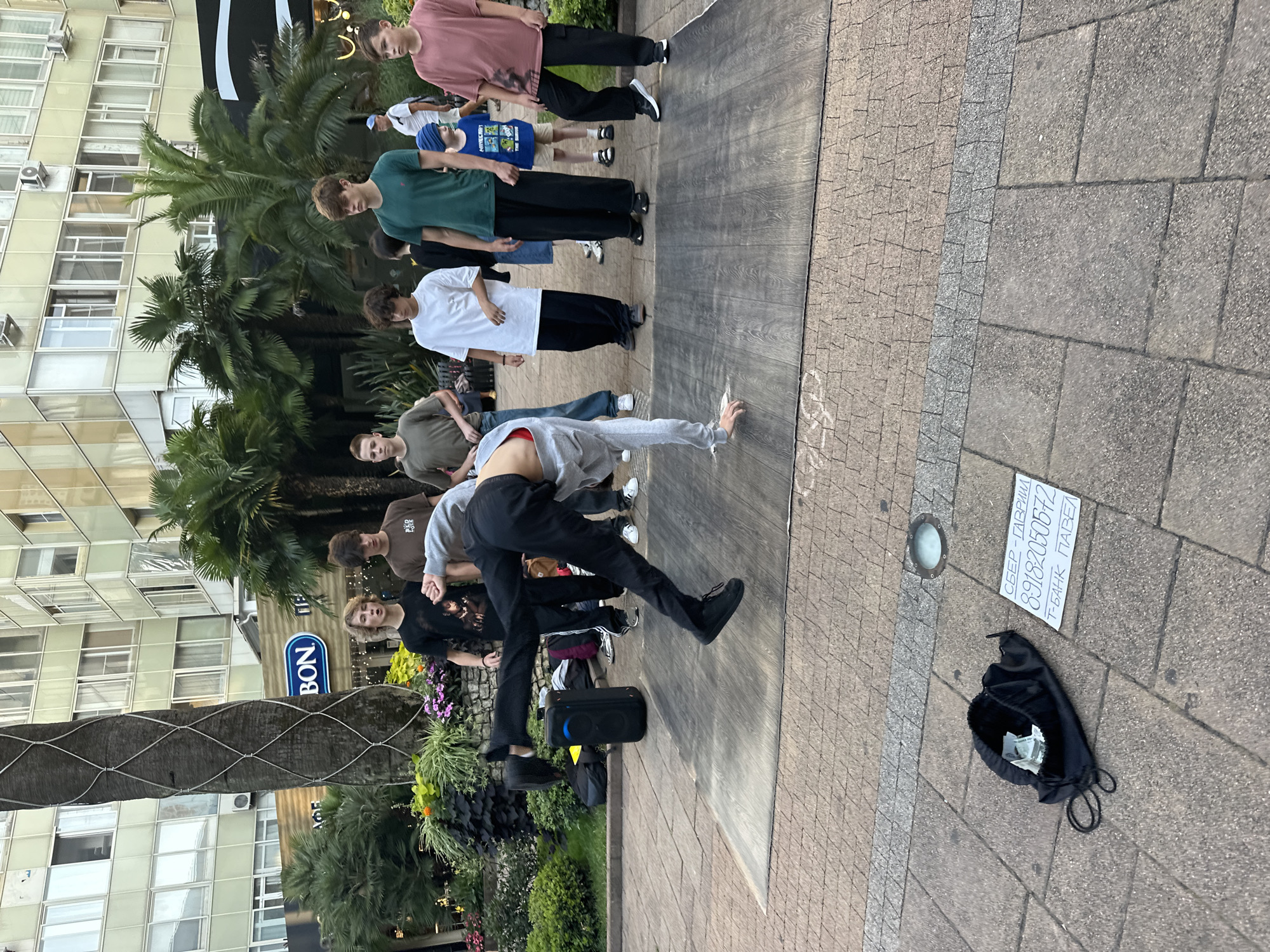 In Sochi, Russia, a breakdancer is captured mid-move on a wooden platform laid on a paved walkway. The dancer, wearing black pants and a grey hooded jacket with a red band visible at the waist, is performing a freeze, with one hand on the ground and legs extended in the air. A black speaker sits to the left of the platform, near a black drawstring bag containing cash and a white sign with Cyrillic text that reads "СБЕР - ГАВРИИЛ 89182050572 Т-БАНК ПАВЕЛ". Several young people, dressed in casual clothing, stand watching the performance. Behind them, lush palm trees and flowering bushes are visible, with a multi-story building featuring numerous windows in the background. The overall scene suggests an outdoor performance or street art event in a public urban space.