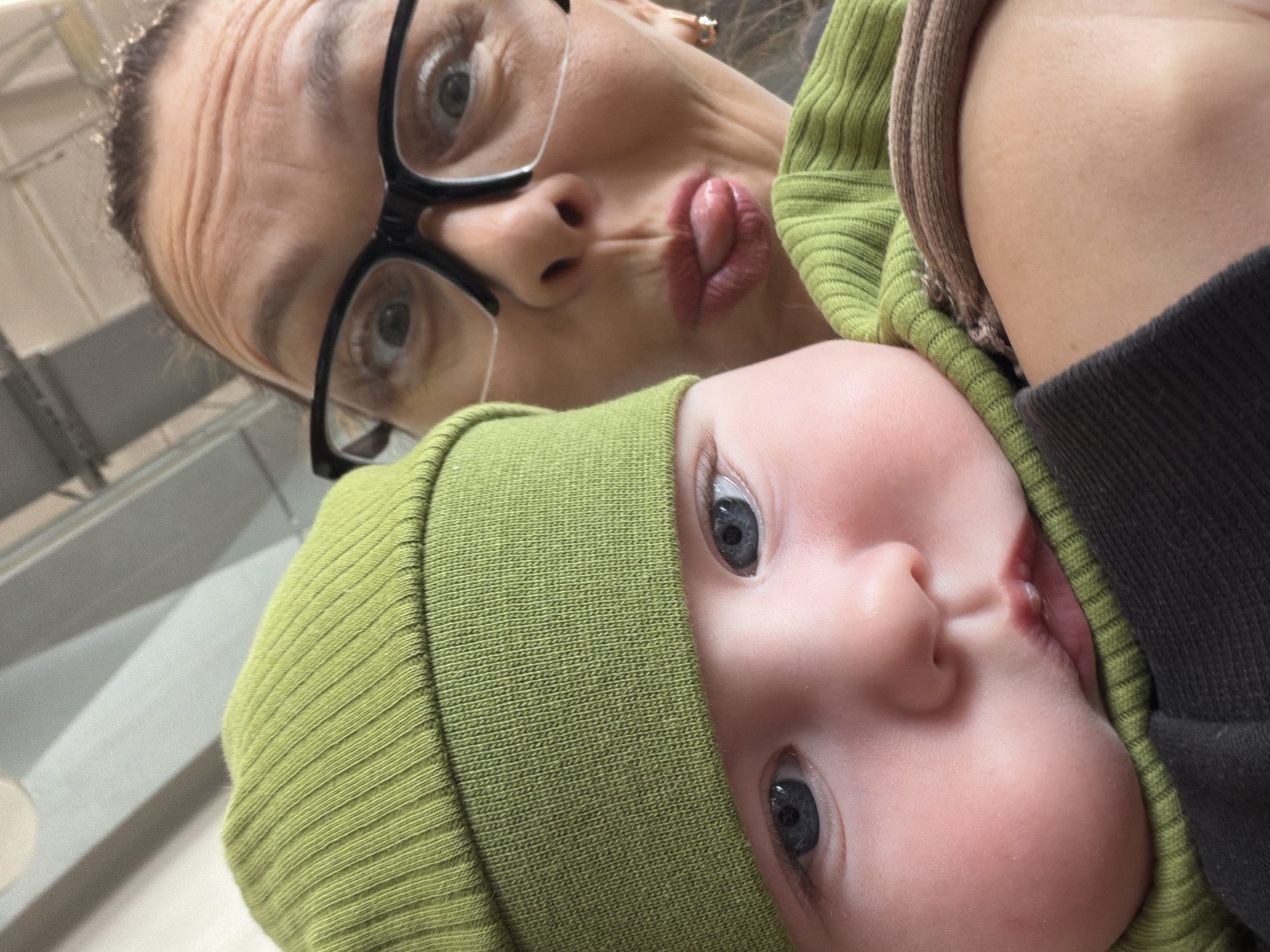 A close-up selfie of a woman and a baby. The woman is wearing black-framed glasses and making a puckered face. The baby is wearing a green knitted hat. They are indoors with a white ceiling visible in the background. There is no location information in this image that indicates it was taken in the city of Chaikovsky, Russia.