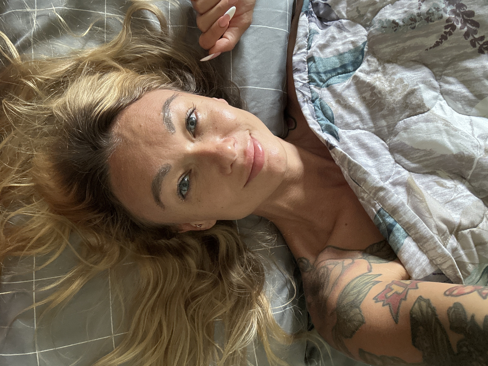 A woman with blonde hair and blue eyes lies on a bed with a grey and white checkered sheet. Her hair is spread out around her head. She is wearing a grey and white patterned comforter that covers her chest and lower body. Tattoos are visible on her left arm and shoulder. Her right hand, with long, almond-shaped nails, is resting on the sheet near her face. The image is a close-up, high-angle shot. The setting does not provide specific clues to indicate a location in Mũi Né, Vietnam.