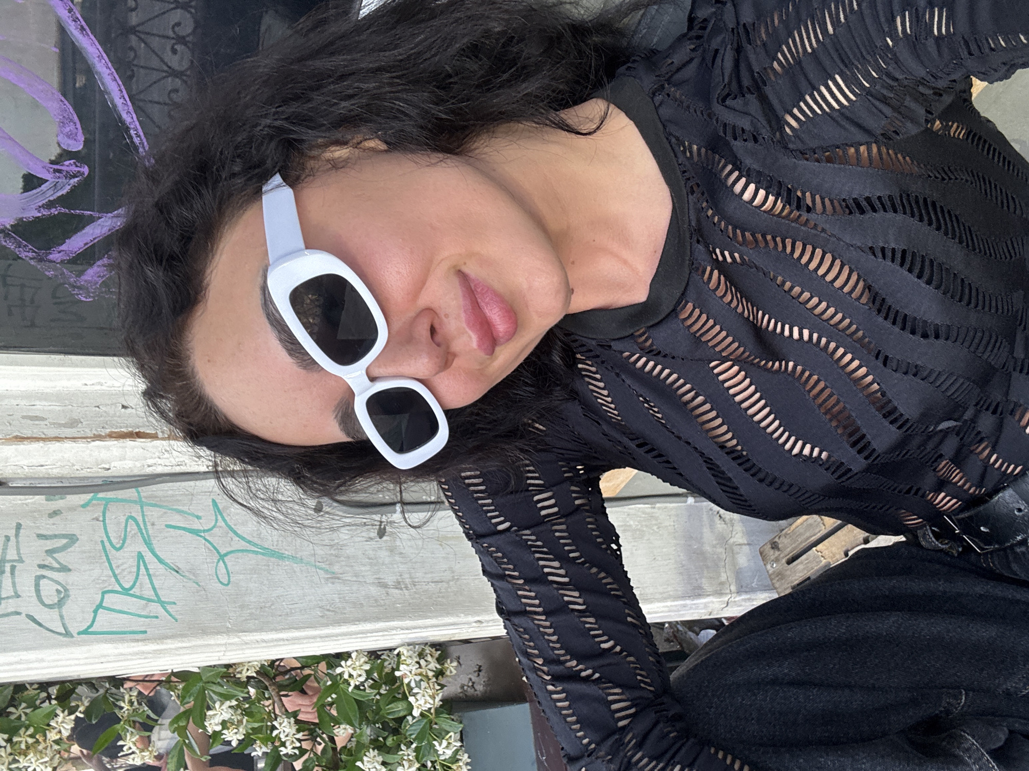 In the city of Tbilisi, Georgia, a woman with dark hair and white-framed sunglasses is captured in a selfie. She is wearing a black, semi-sheer top with an intricate pattern of cutouts. The background features a concrete wall with graffiti, including teal markings and purple streaks. To the left, lush green foliage with small white flowers is visible, suggesting an outdoor setting. The woman is seated, with her left arm resting on her thigh and her body angled towards the camera.