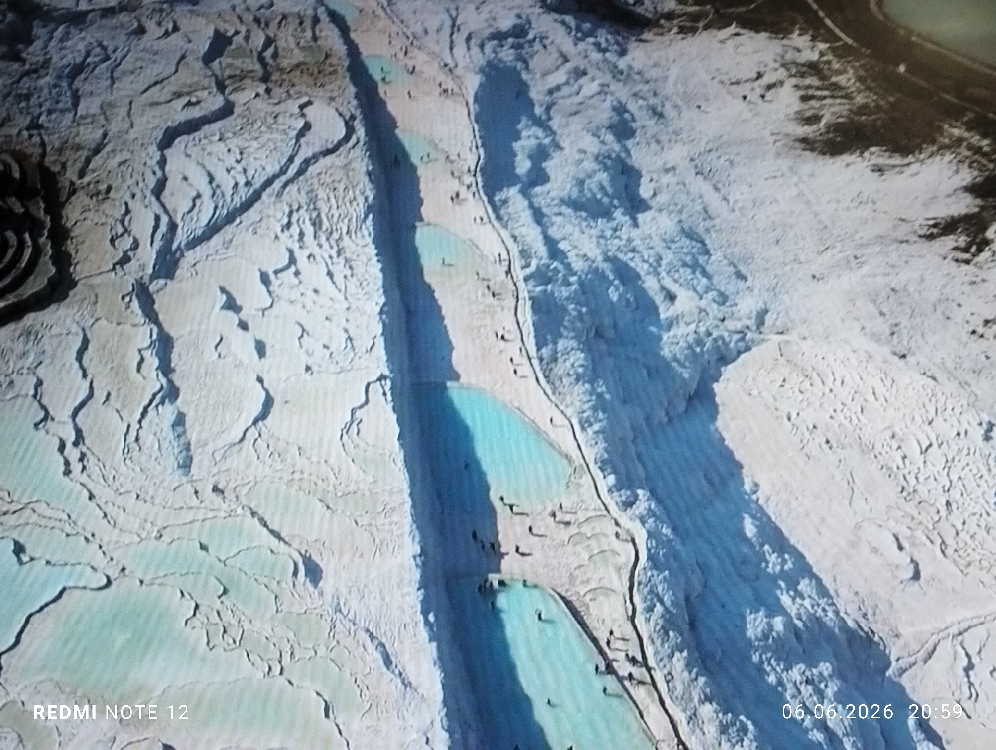 This image presents an aerial or high-angle view of the Pamukkale travertine terraces in Turkey. The scene is dominated by vast formations of white calcium carbonate, sculpted into intricate terraced pools and cascades. Numerous natural basins hold strikingly vivid light blue to turquoise thermal water.
A prominent, elongated channel or pathway, appearing darker and deeper, bisects a significant portion of the landscape from the upper center towards the lower right. Along and within this central feature, as well as in the surrounding natural pools, are numerous small, dark figures, indicating the presence of people wading, bathing, or walking. These individuals are dispersed across the accessible areas of the terraces.
On the far left, the white formations appear more fragmented and spread out, forming a mosaic of smaller, shallower pools. The right side of the image features a steeper, more densely packed arrangement of the white terraces. Towards the upper right corner, the terrain transitions to a darker, possibly drier or shadowed area.
The overall environment suggests a natural geological wonder, heavily frequented by visitors interacting with the unique thermal pools. Text overlays in the bottom left corner read "REDMI NOTE 12," and in the bottom right corner, "06.06.2026 20:59" displays a date and time.