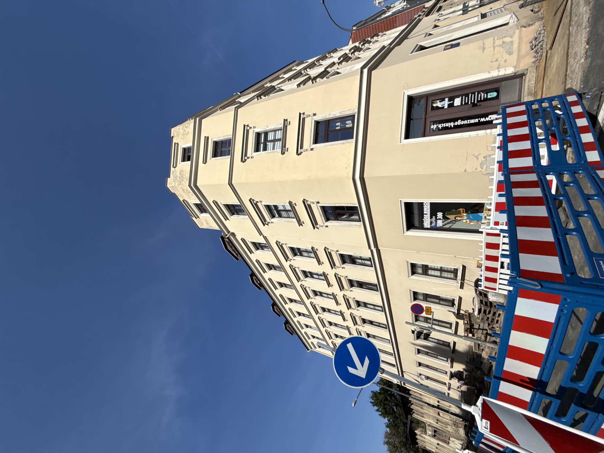The image depicts a corner of a pale yellow building in Görlitz, Germany, under a clear blue sky. The building has multiple stories with numerous windows, some featuring decorative frames. Construction barriers, consisting of blue and white plastic fences with red and white striped panels, are visible in the foreground, indicating ongoing work. A blue and white circular traffic sign with a white arrow pointing left is mounted on a pole. A traffic light and a circular red and white sign are also present. Several pallets are stacked near the building, and loose stones are scattered on the ground. A store window displays an advertisement featuring a man carrying a box and text that reads "UMZÜGE BINSCH" and a phone number. Another door has text and a website address: "www.umzuege-binsch.de". The overall scene suggests a street view in a city area undergoing construction.
