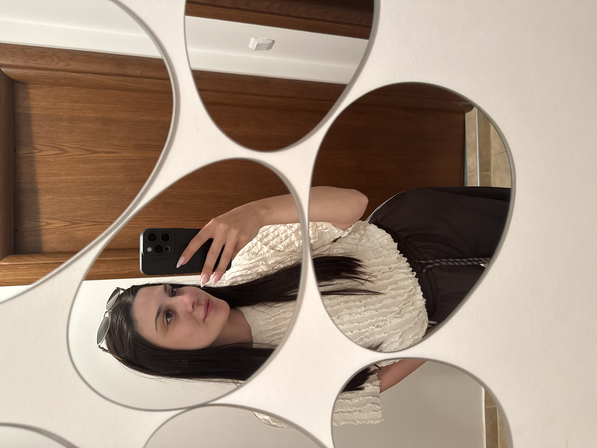 A young woman is captured in a mirror selfie, with the image rotated 90 degrees clockwise. She appears in the reflection, holding a dark-colored smartphone with her right hand to take the picture. Her dark hair is long and straight, with sunglasses perched on top of her head. She wears a distinctive white, ruffled long-sleeved top and dark-colored pants or a skirt with a visible drawstring detail. Her nails are manicured with a light design.
The mirror she is using has a unique, multi-segmented design with white borders, fragmenting the reflection into several irregular shapes. Through these mirrored sections, elements of an indoor setting are visible. These include warm, brown wooden paneling, which could be part of a door or wall, and sections of white wall or ceiling. A small, square white light fixture is reflected in one of the upper mirror segments. Another smaller segment at the bottom right hints at a tiled surface, suggesting the location might be a bathroom or a room with tiled features. The woman's expression is neutral as she looks towards the camera. No text is visible in the image.