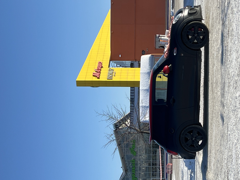 The image displays a black matte Mini Cooper car with a red roof and red side mirrors, parked in a snow-covered outdoor retail parking area. Several car cleaning items, including multiple spray bottles, a white bucket, and cloths, are positioned on the vehicle's hood and windshield. A single individual, dressed in dark clothing, is partially visible near the front right fender of the car, seemingly engaged in or preparing for car cleaning. The vehicle is equipped with black multi-spoke wheels and winter tires.
In the background, under a clear blue sky, two commercial buildings are prominent. To the left, a large glass-fronted building displays green Cyrillic lettering "О’КЕЙ ДОМ" and "Садовый Рай," identifying it as an O'KEY Dom hypermarket and garden center. To the right, a multi-story structure features a distinctive yellow, angular facade with red Cyrillic lettering "М.Видео" (M.Video) and "ВВВВ," indicating an electronics retailer. This yellow structure is connected to an adjacent orange building. Bare deciduous trees are visible between the two main background structures. Snow covers the ground and some building ledges. White and red barrier tape is present in the distant left background. The specific location is identified as a retail complex at Pulkovskoye Shosse 17 к2, Saint Petersburg, Russia.