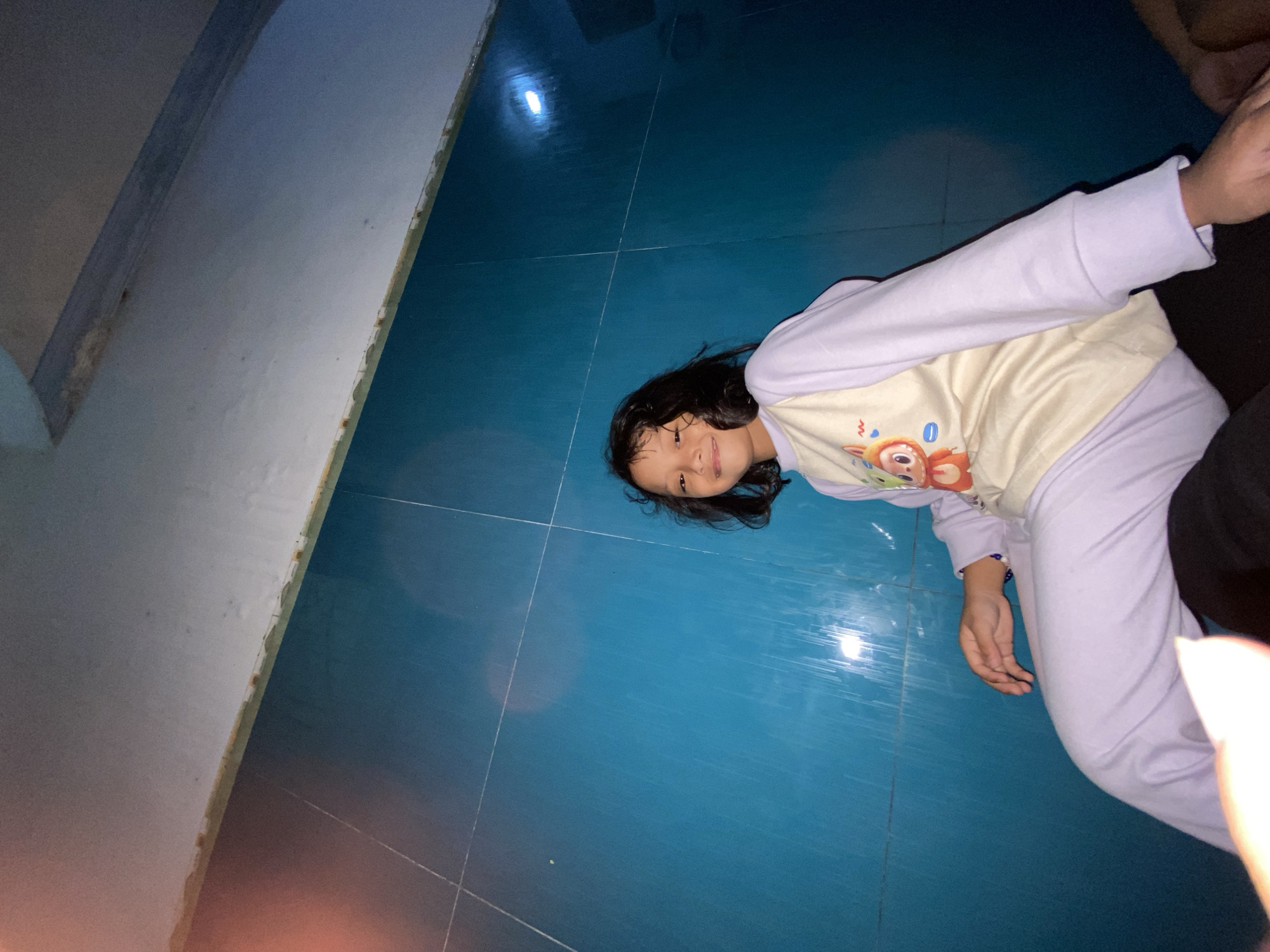 This image, captured in Gelam, Indonesia, depicts a young girl smiling and looking directly at the camera. She has shoulder-length dark hair and is wearing light purple long-sleeved pajamas with an off-white front featuring a cartoon graphic of an orange, large-eyed animal character surrounded by small blue and orange shapes. Her left hand, adorned with a blue beaded bracelet, rests on her upper thigh. She is seated on a dark surface, possibly another person's lap, partially visible at the bottom of the frame. The background consists of a prominent bright blue tiled wall, displaying distinct grid lines and vertical streaks, suggesting dampness or surface residue. Above the blue tiles, a lighter, possibly off-white or light grey, wall or ceiling section is visible, separated by an unfinished or irregular border. Artificial lighting creates reflections on the blue tiled surface, with a notable bright spot in the upper right. A subtle orange lens flare is present in the upper left portion of the image.